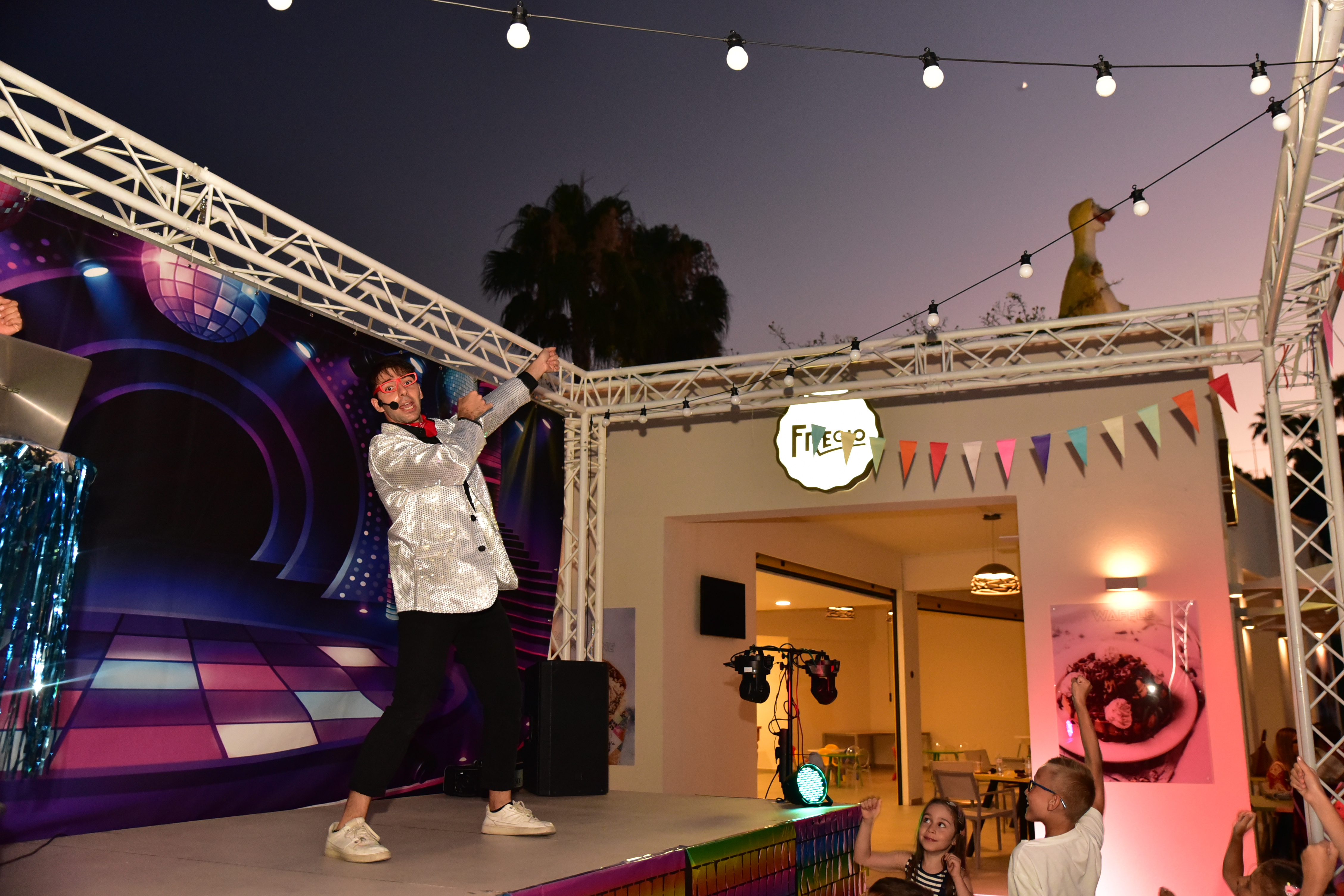 Performer in silver jacket entertaining children on outdoor stage near Fregio cafe during evening show
