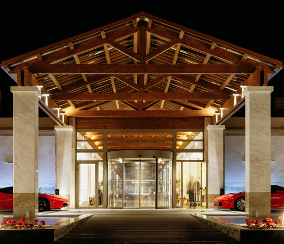 Illuminated main entrance with wooden roof, marble columns, and luxury cars at night