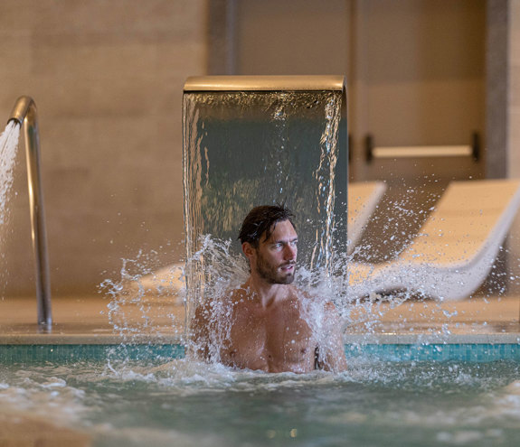 Man enjoying neck massage jet under water cascade at indoor thermal spa pool
