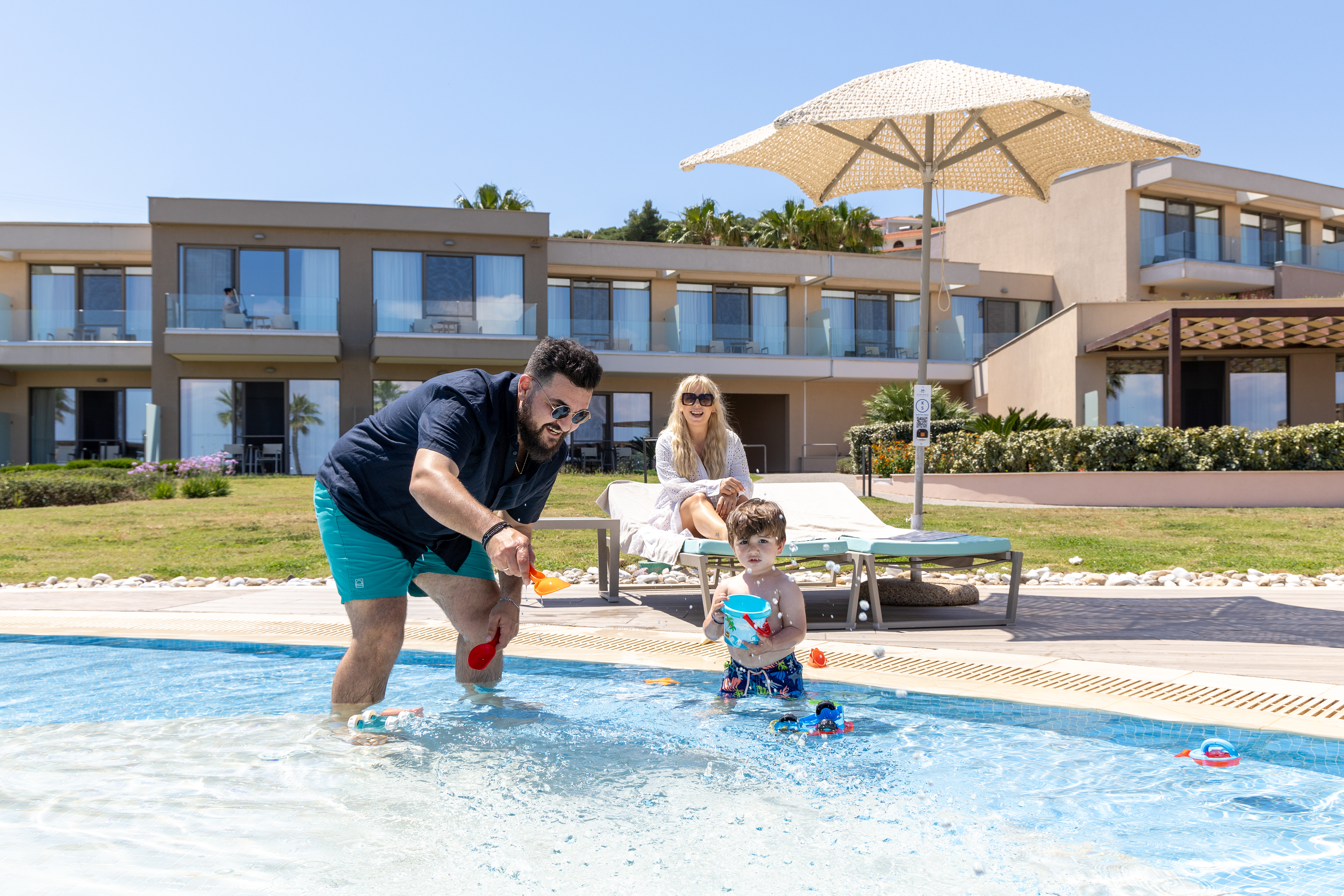 Father playing in the kids' pool with his toddler while mother relaxes on a sunbed under the sun