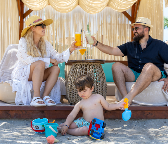 Parents enjoying cocktails in a private beach cabana while their child plays with toys in the end