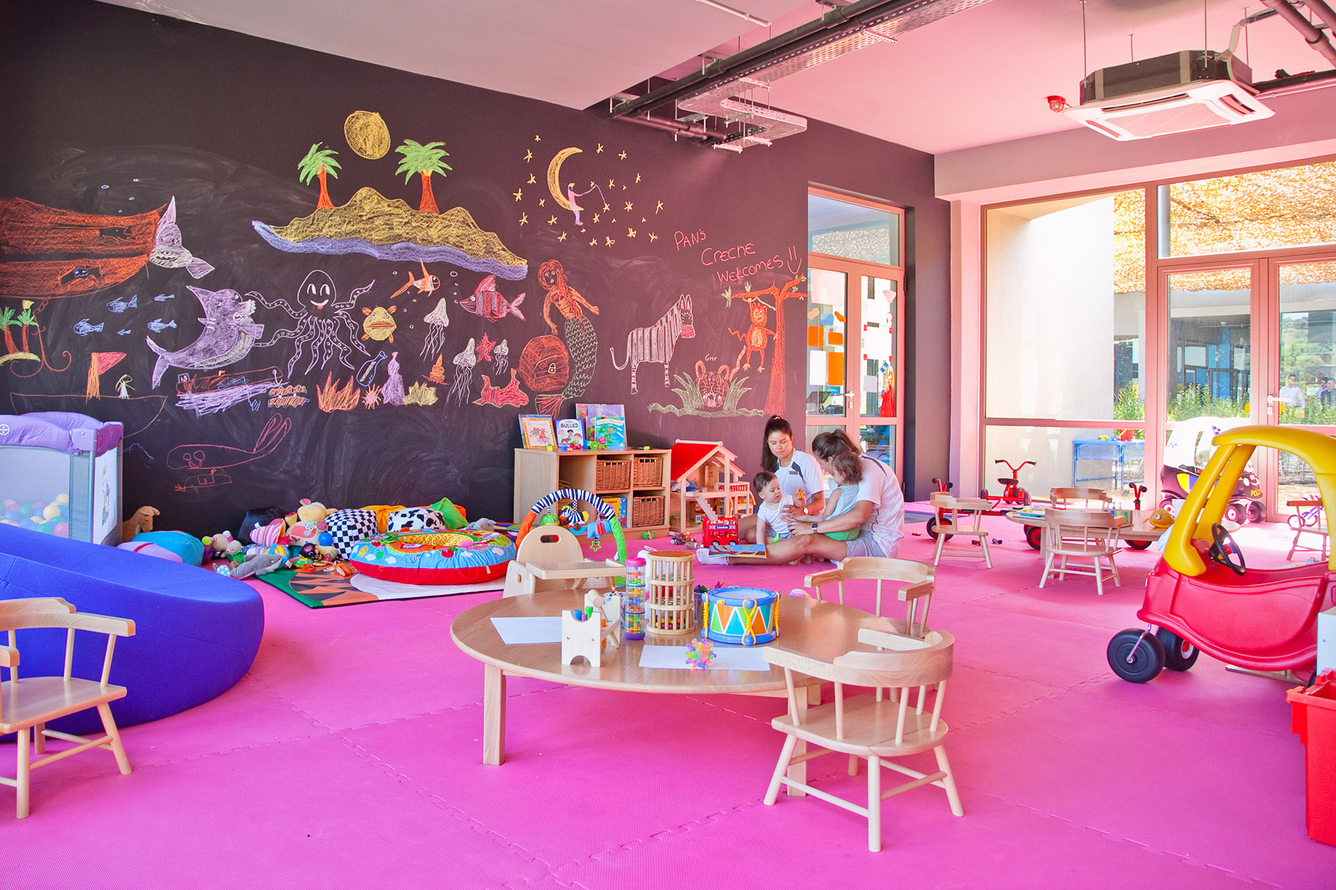Spacious indoor play area in Kids Planet with staff playing with toddler, drawing wall, and toys
