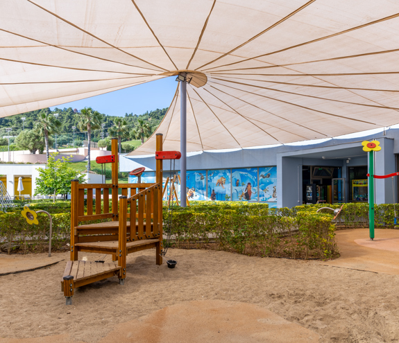 Outdoor shaded playground with wooden climbing structure and sand area at Kids Planet