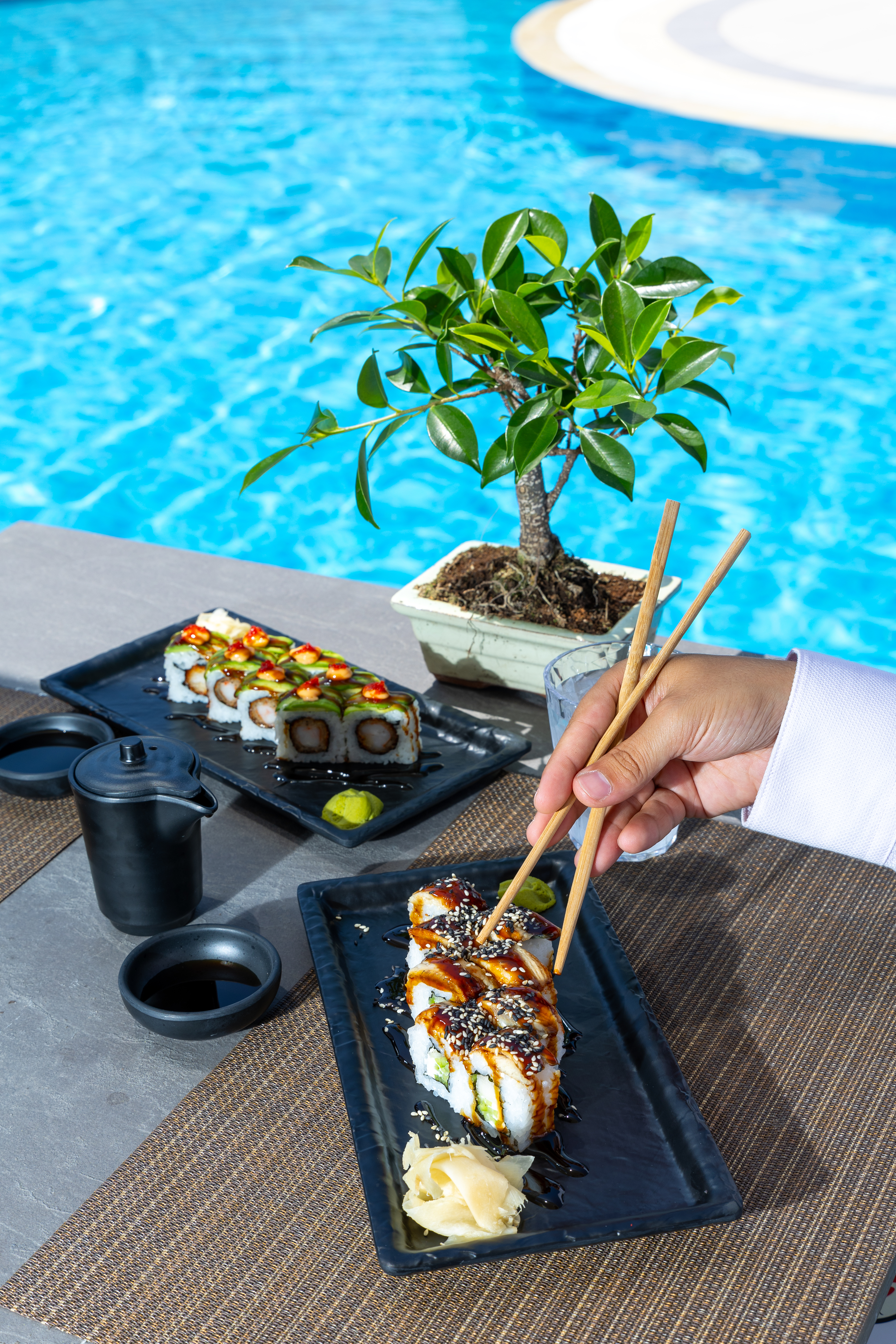 Guest using chopsticks to pick sushi beside a bonsai tree and blue pool water