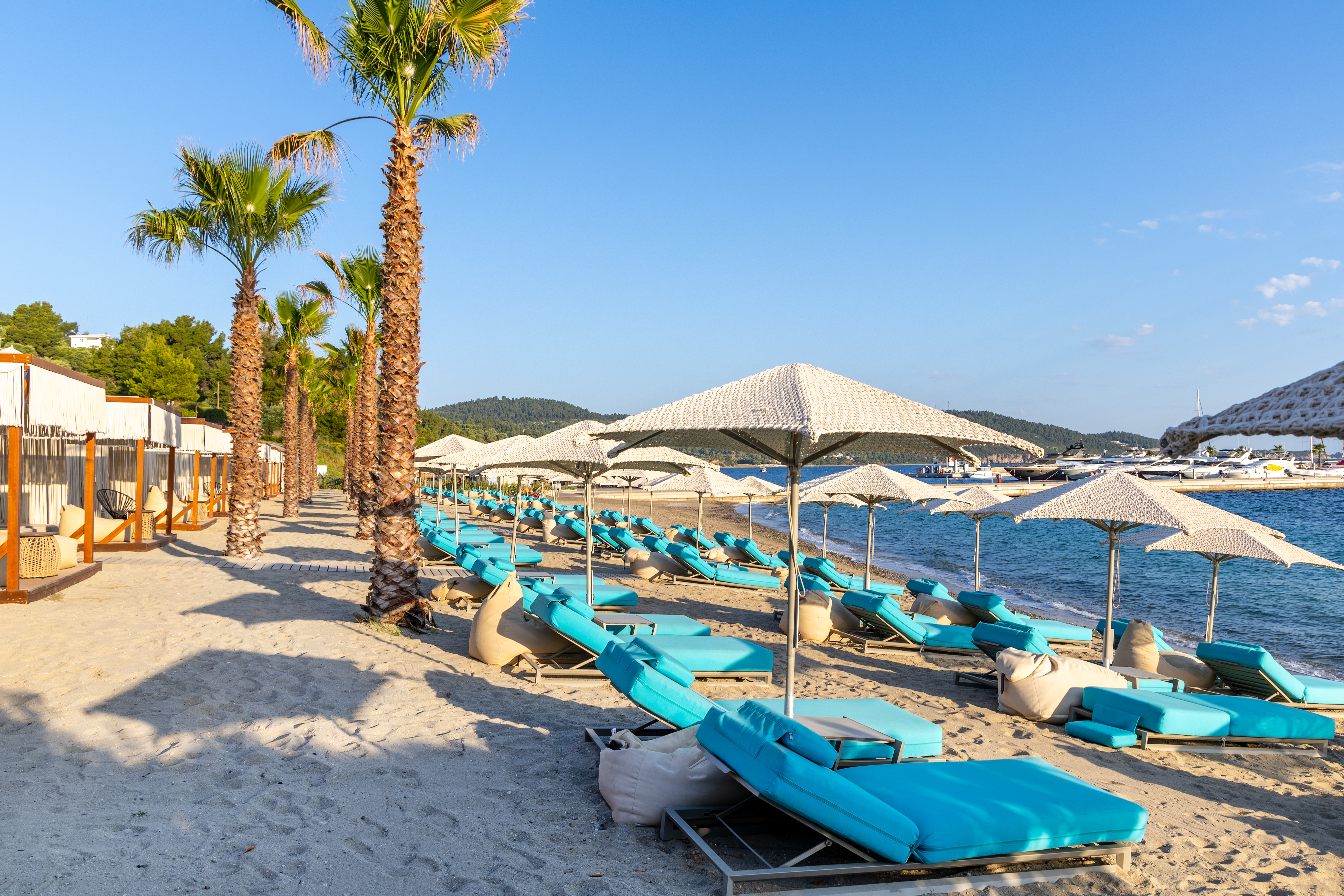 Sandy beachfront lined with palm trees, sunbeds, and woven umbrellas overlooking the calm sea