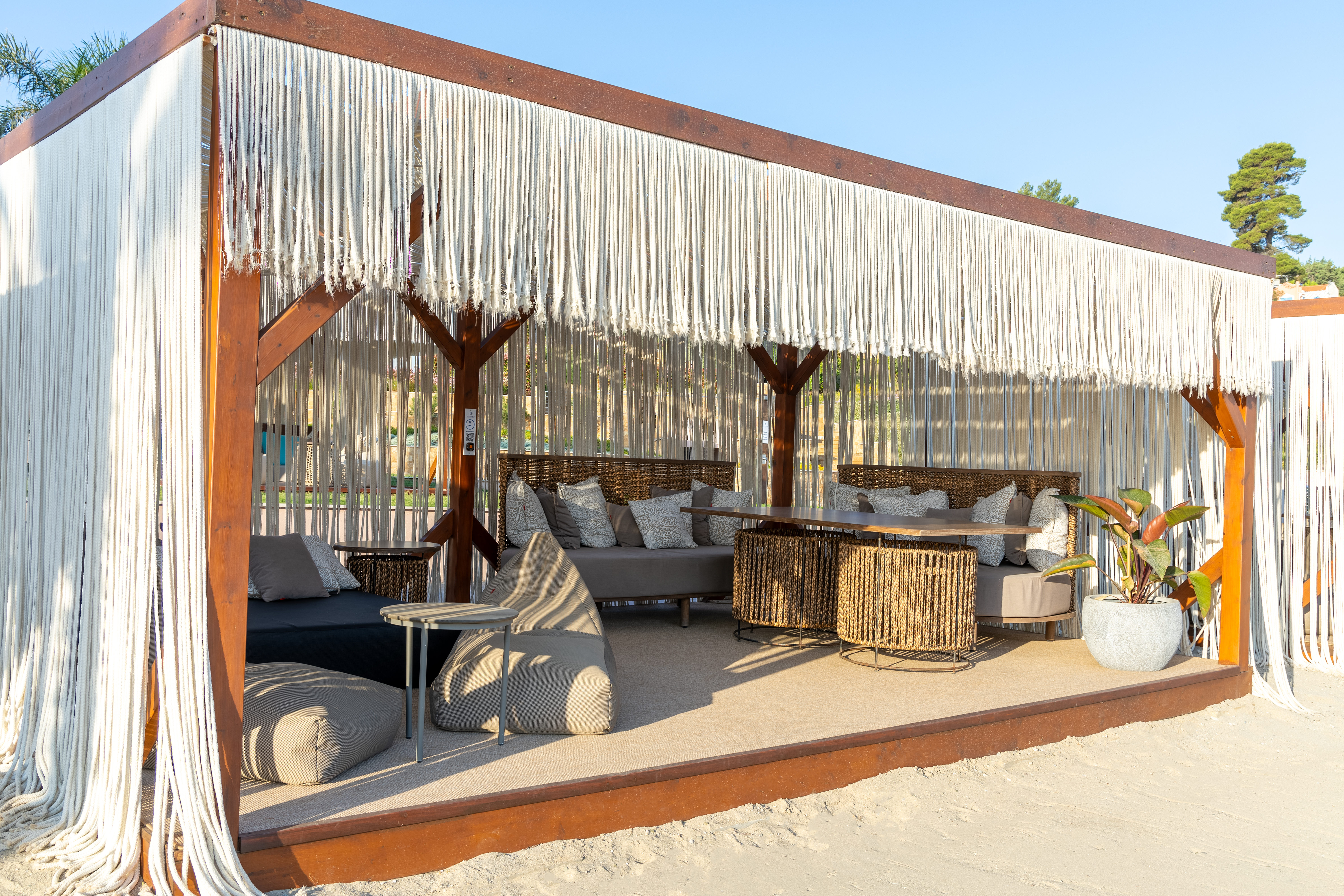 Spacious beach cabana with woven furniture, neutral cushions, and flowing rope curtains on the sand