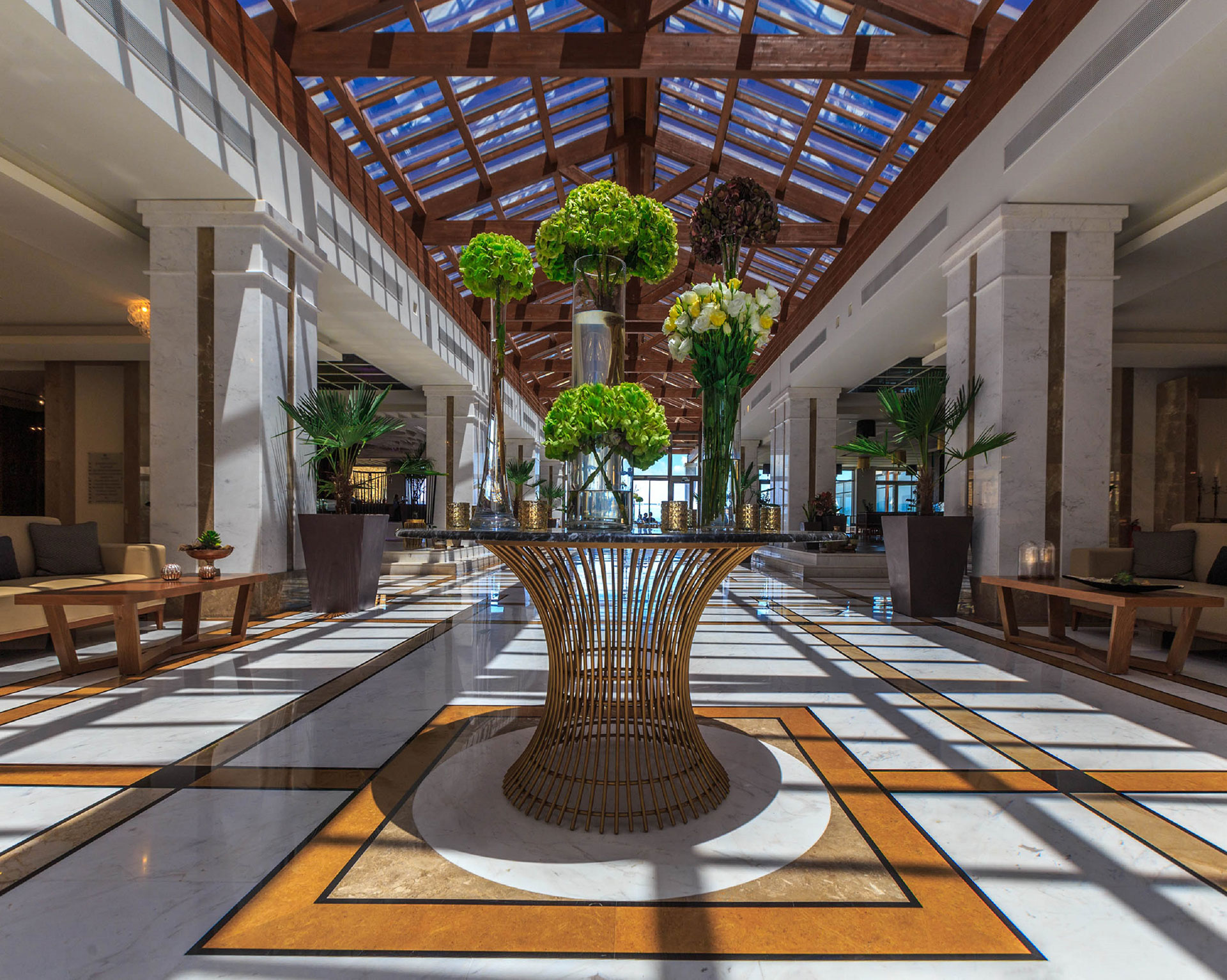 Elegant hotel lobby with marble floors, glass roof, and floral centerpiece bathed in natural light