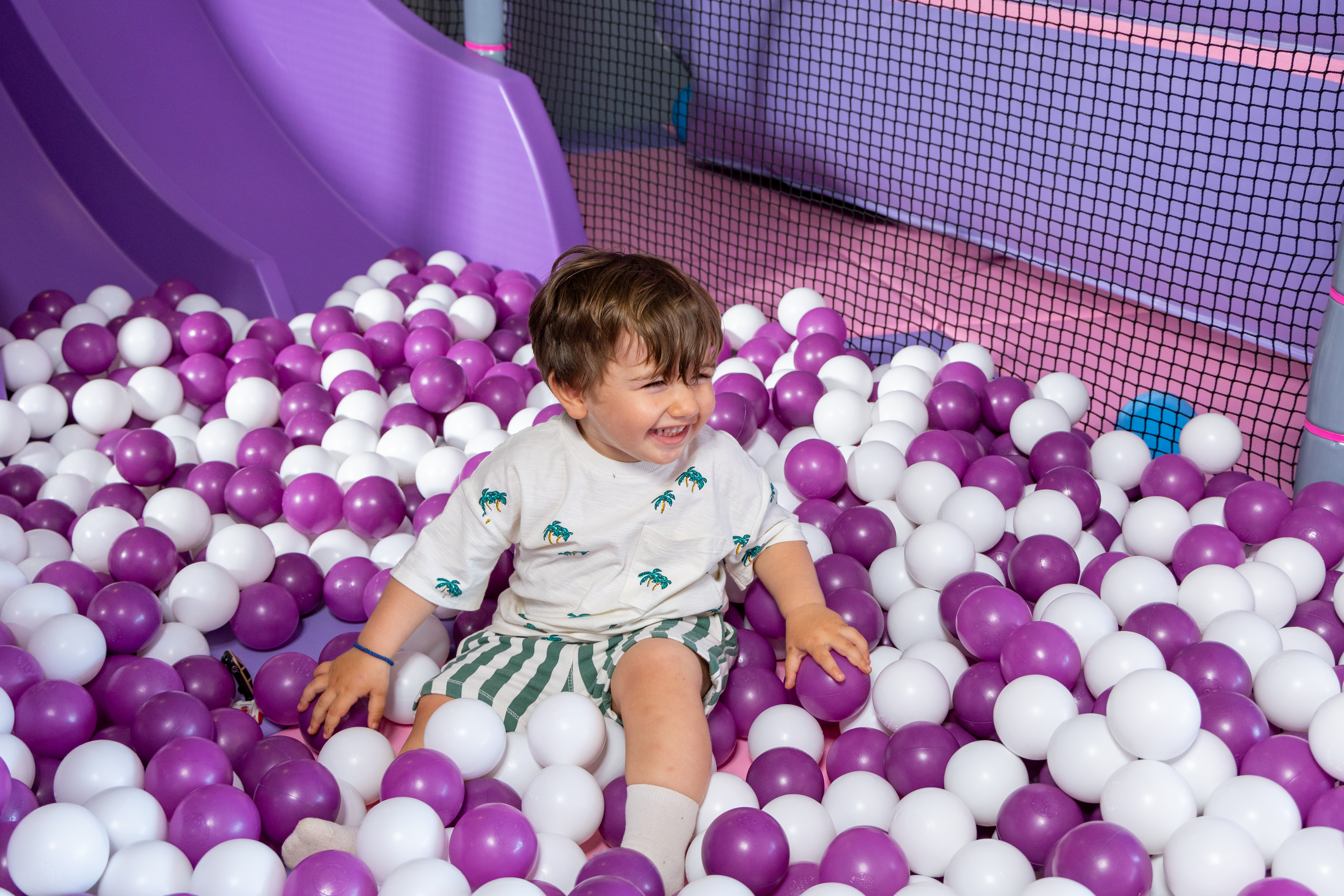 Laughing toddler sitting in a colorful purple and white ball pit in Kids Planet
