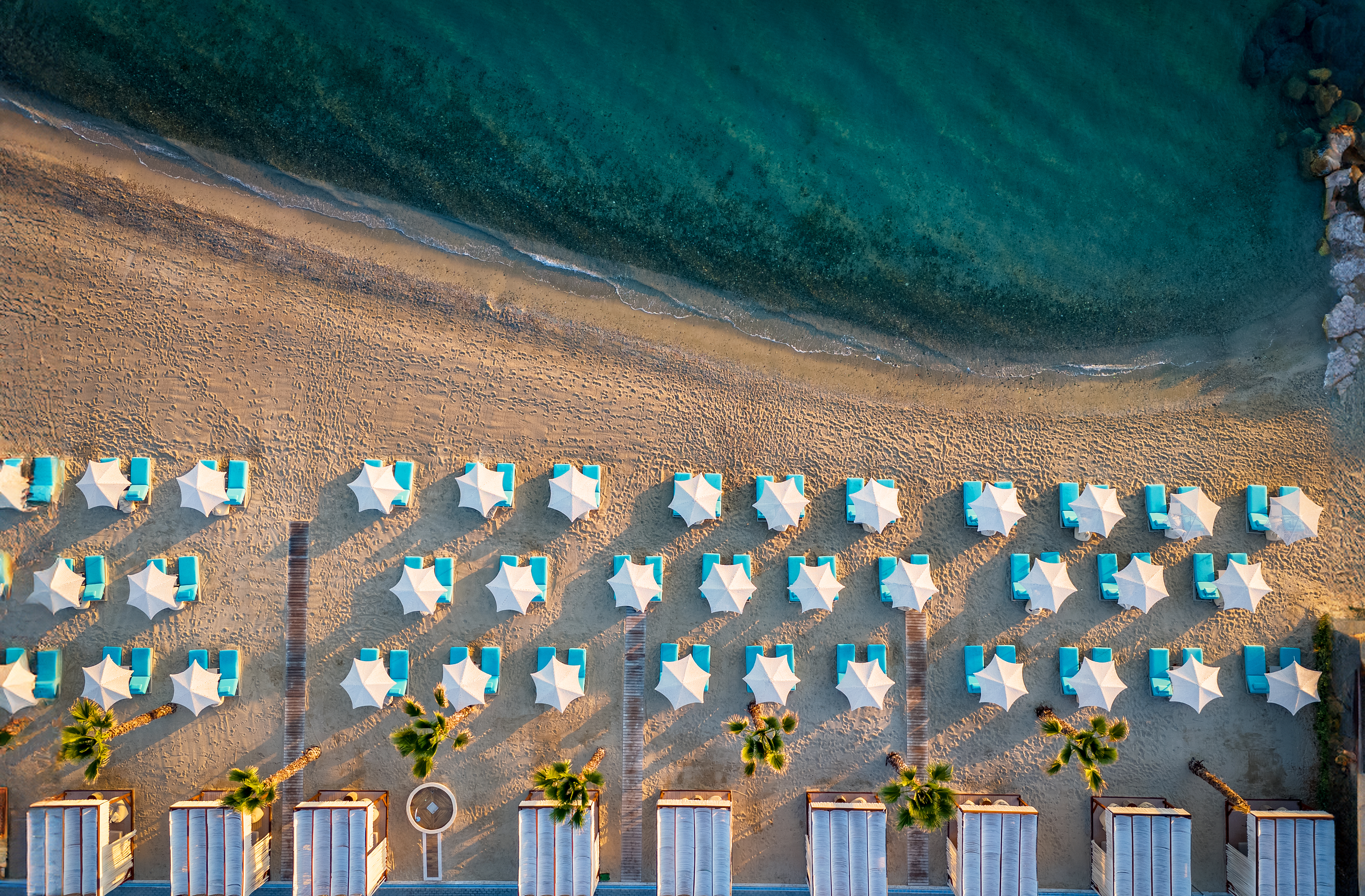 Aerial photo of the sandy beach with turquoise sunbeds, white umbrellas, and calm emerald waters