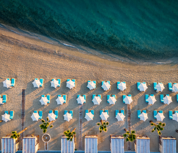 Aerial photo of the sandy beach with turquoise sunbeds, white umbrellas, and calm emerald waters
