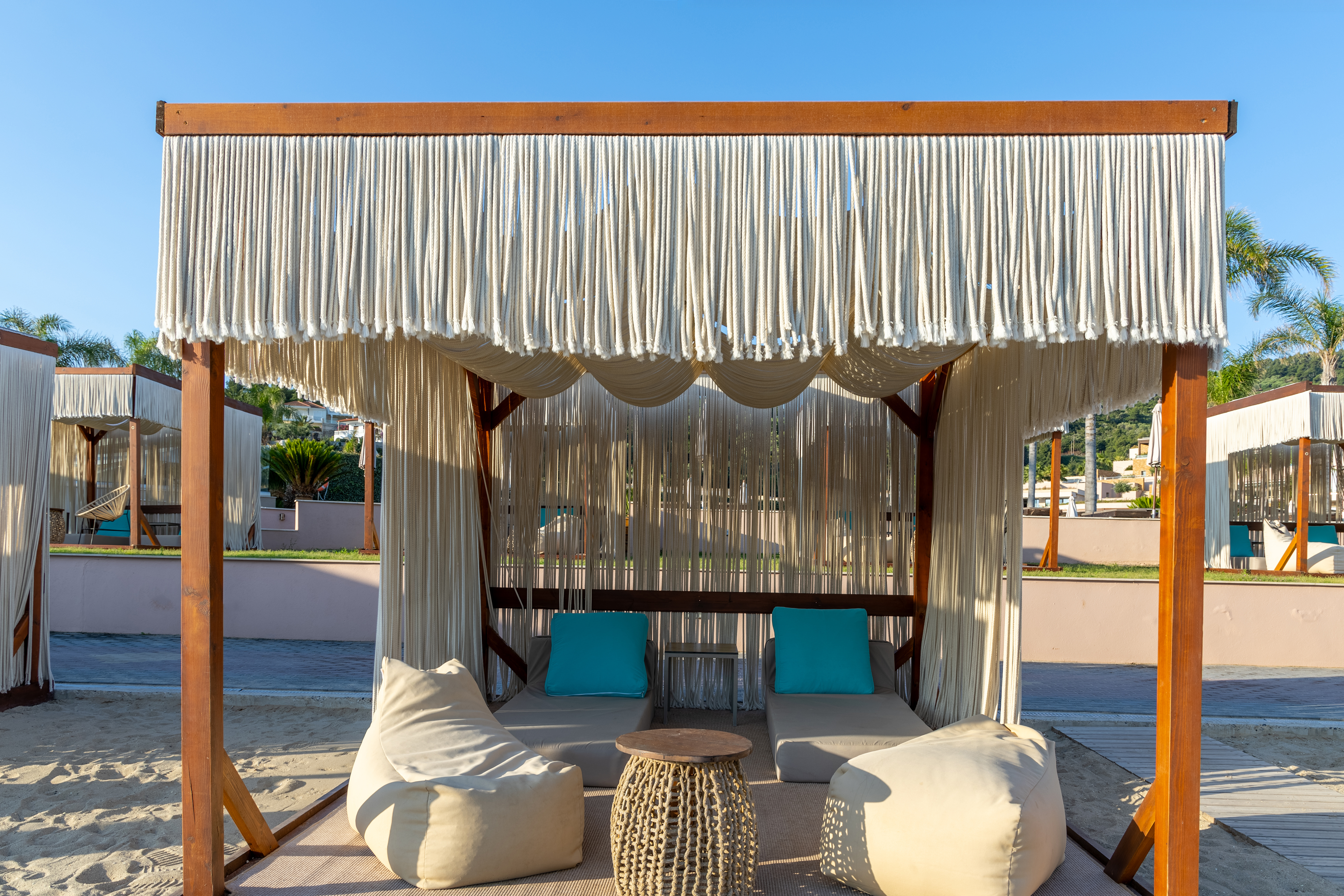 Bohemian style cabana with rope curtains, soft beige loungers, turquoise cushions and wicker table