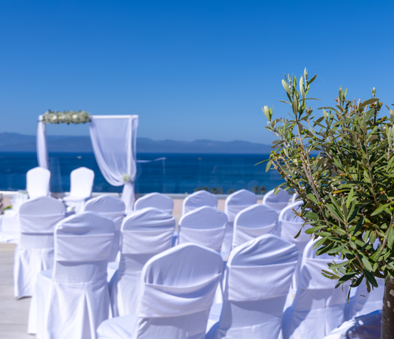 Olive tree and white wedding chairs overlooking the Aegean Sea on a sunny terrace