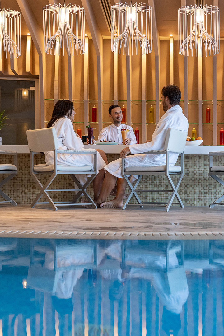 Guests in robes enjoying drinks by the indoor spa bar beside the pool