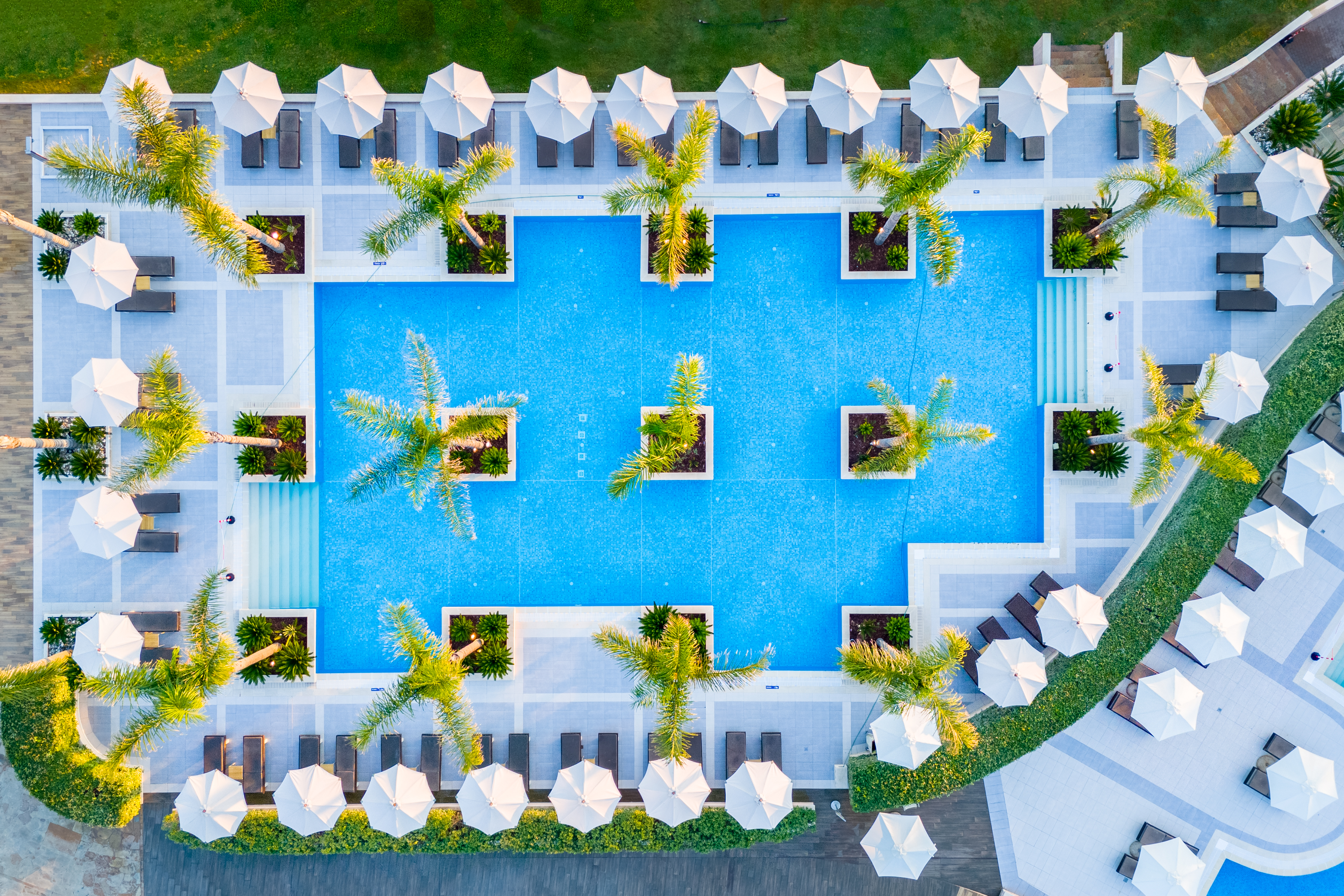 Aerial top view of the geometric pool area with palm trees, white umbrellas, and sunbeds at Mirragio Resort