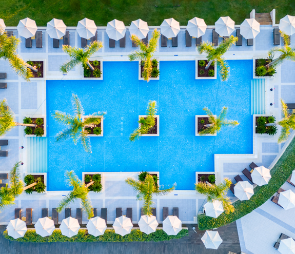 Aerial top view of the geometric pool area with palm trees, white umbrellas, and sunbeds at Mirragio Resort