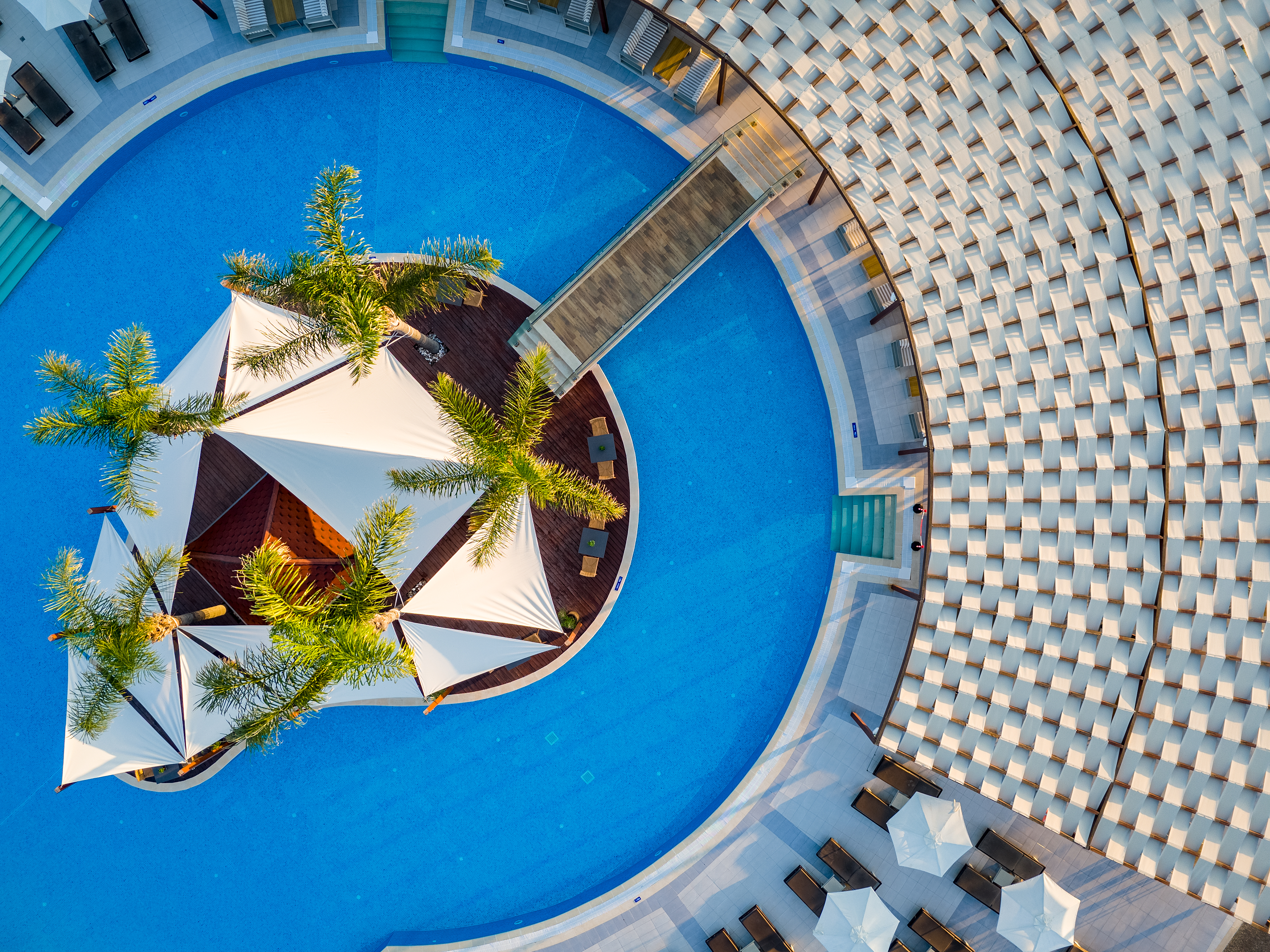 Aerial close up of the resorts central pool with white canopies, palm trees, and sun loungers arranged in perfect symmetry