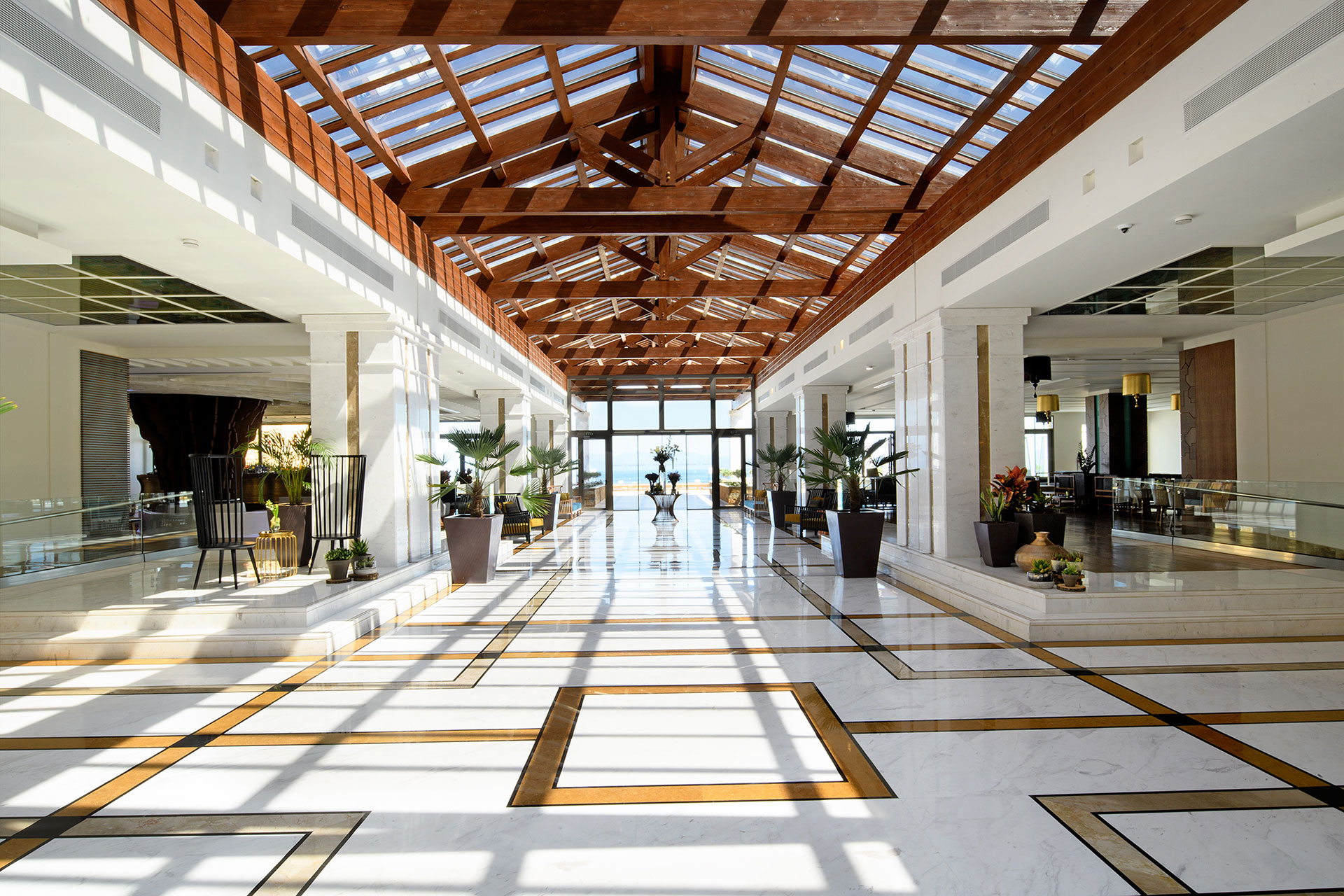 Spacious hotel lobby with marble floors, wooden glass roof, and potted plants offering sea view