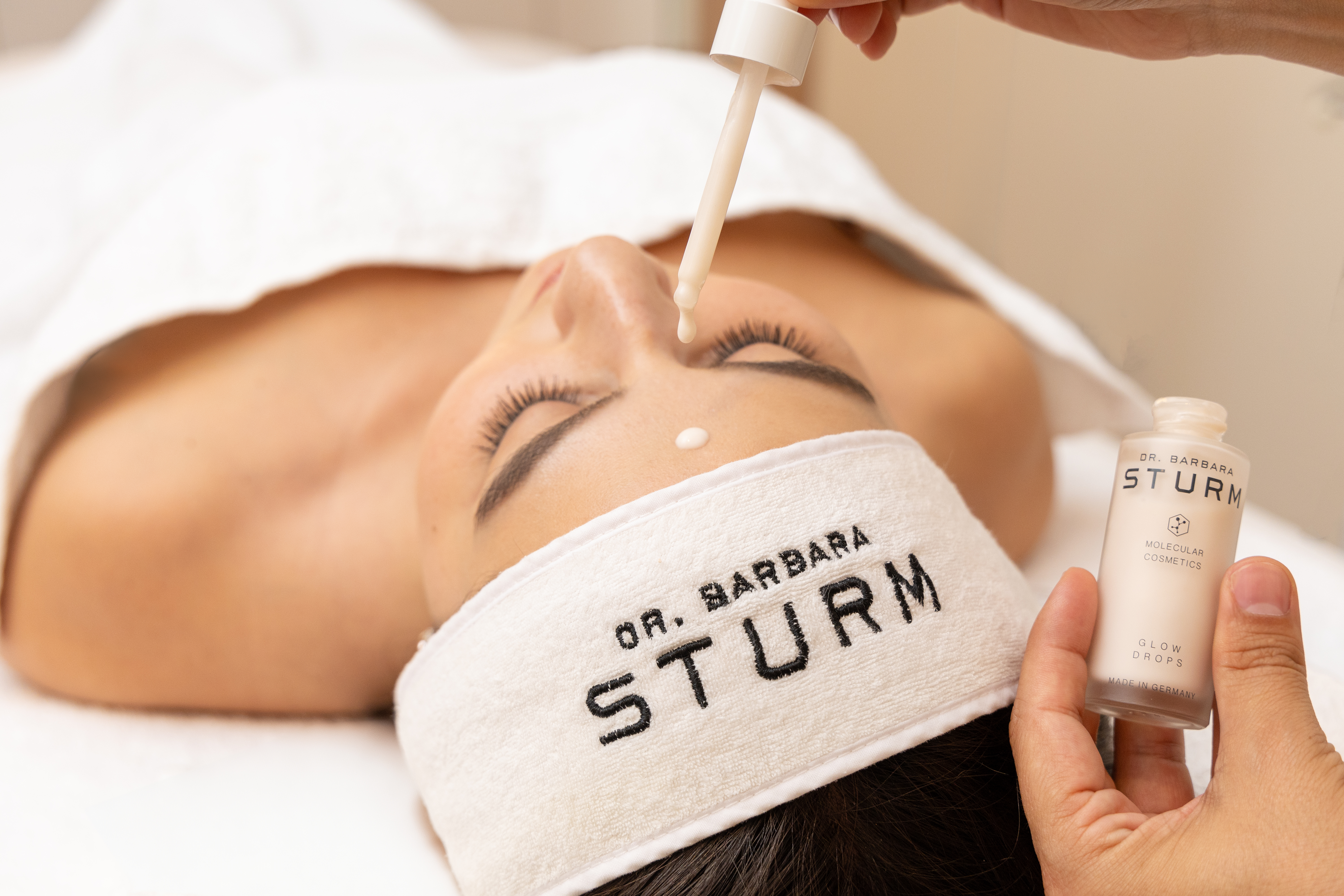 Woman relaxing in modern treatment room while therapist applies Dr Barbara Sturm skincare products to her face
