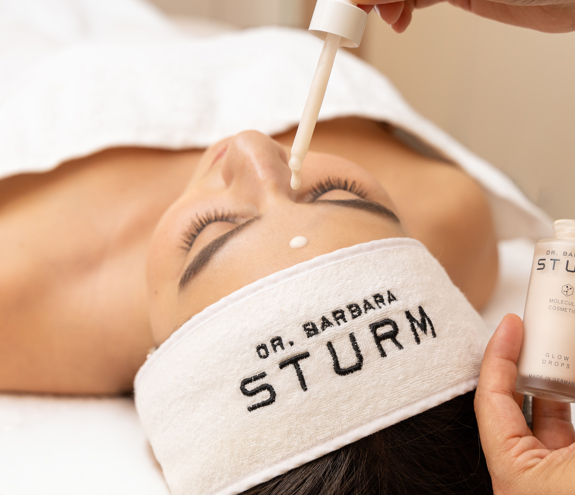 Woman relaxing in modern treatment room while therapist applies Dr Barbara Sturm skincare products to her face