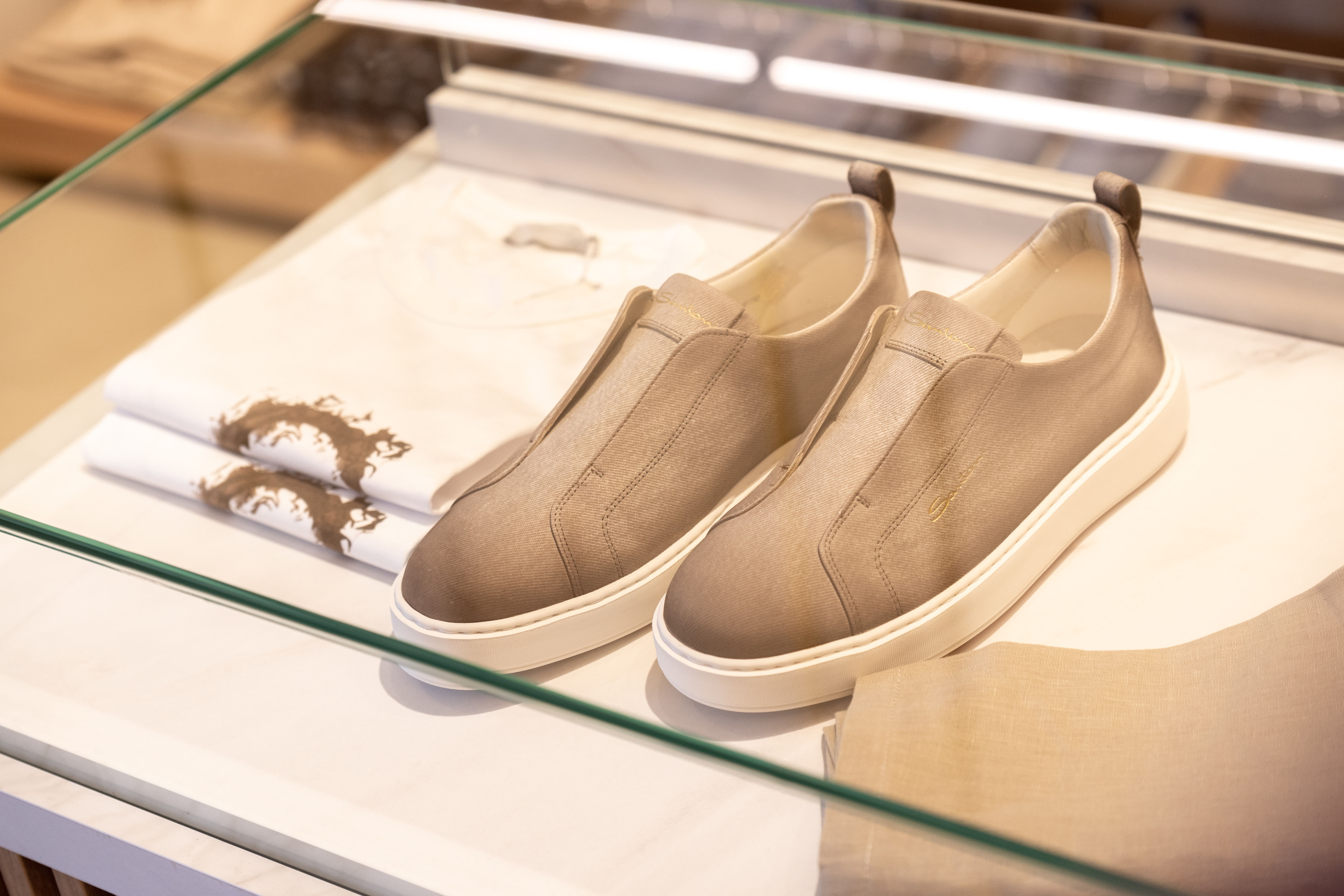 Beige slip on sneakers displayed on glass shelf beside folded white shirts