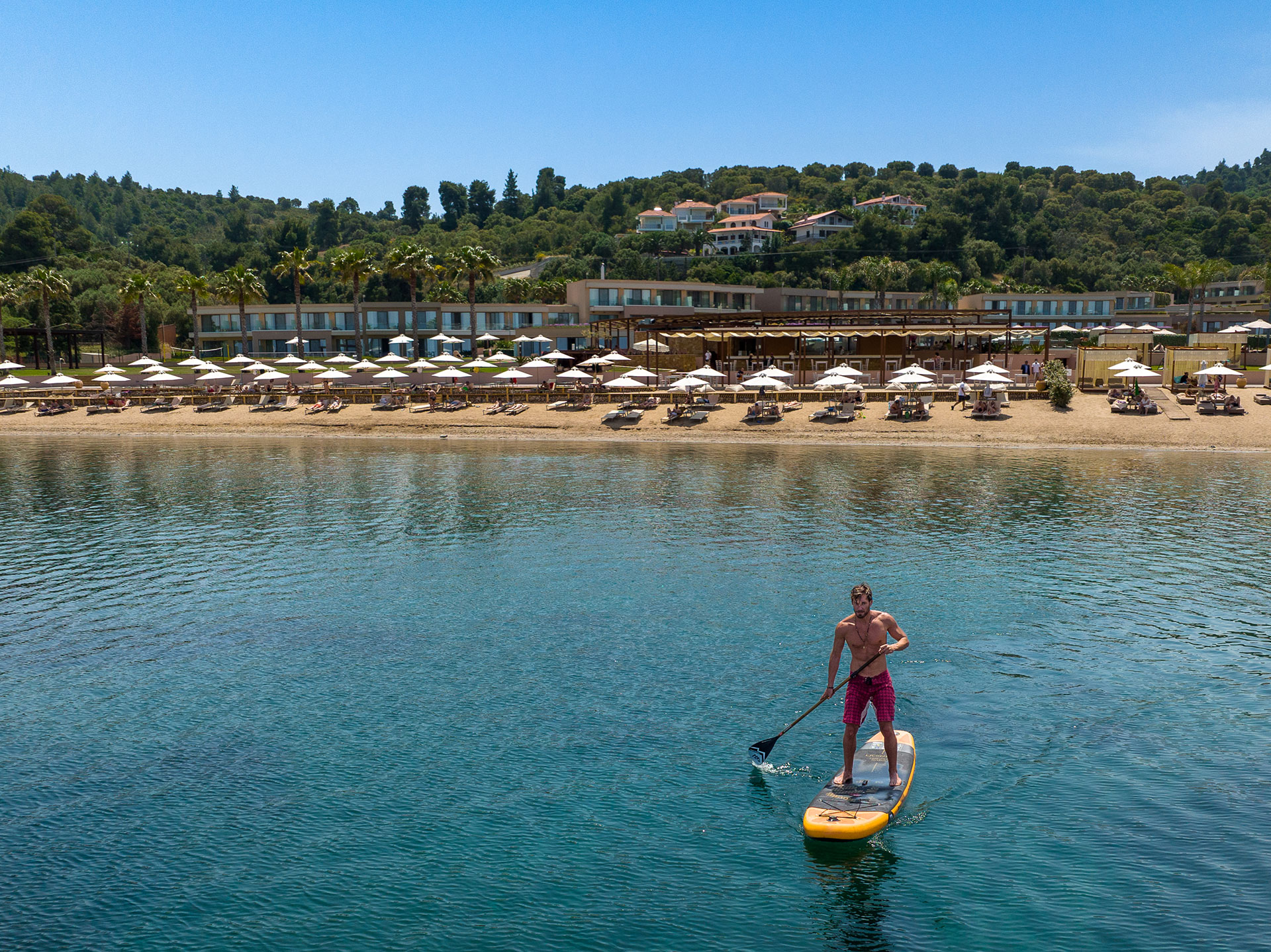 Man paddleboarding on calm turquoise water in front of a sandy beach with sunbeds and umbrellas