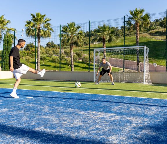 Man kicking football towards goal on outdoor court surrounded by palm trees