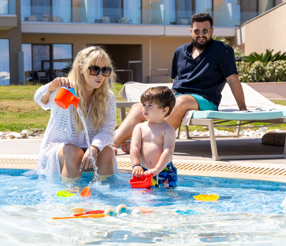 Mother playing with her toddler in the kids' pool while father relaxes on a sunbed nearby