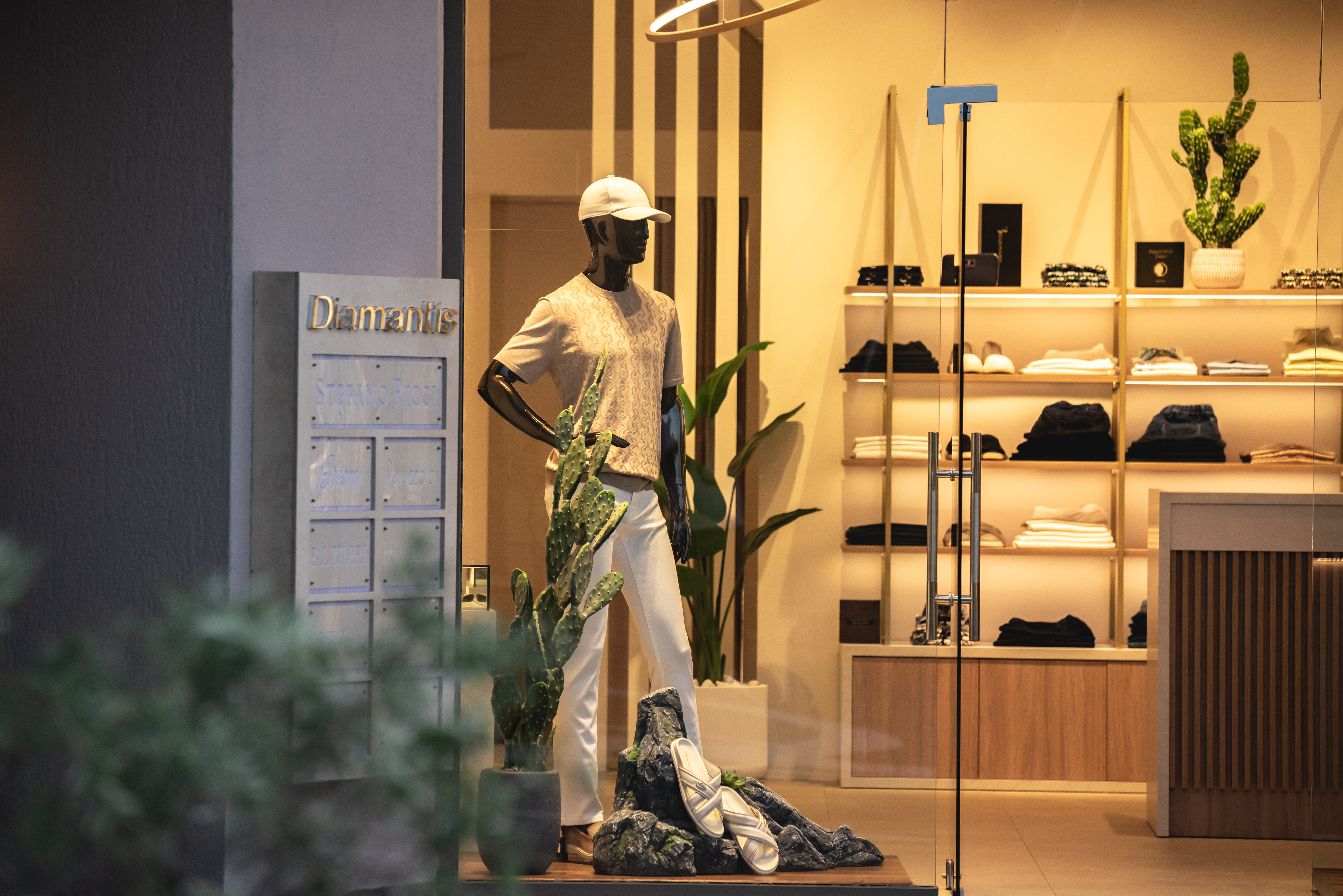 Close up of Diamantis boutique window display with men's casual outfits, cactus decor, and elegant interior lighting
