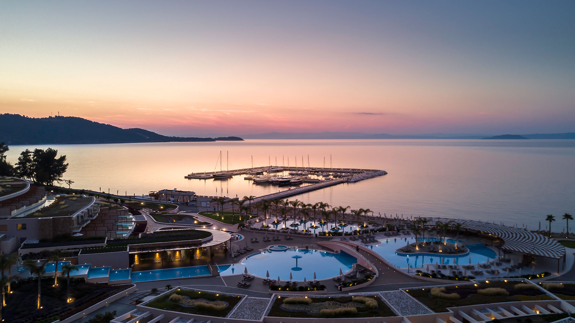 Aerial view of the marina and Miraggio resort's pools at sunset with calm sea and pastel sky