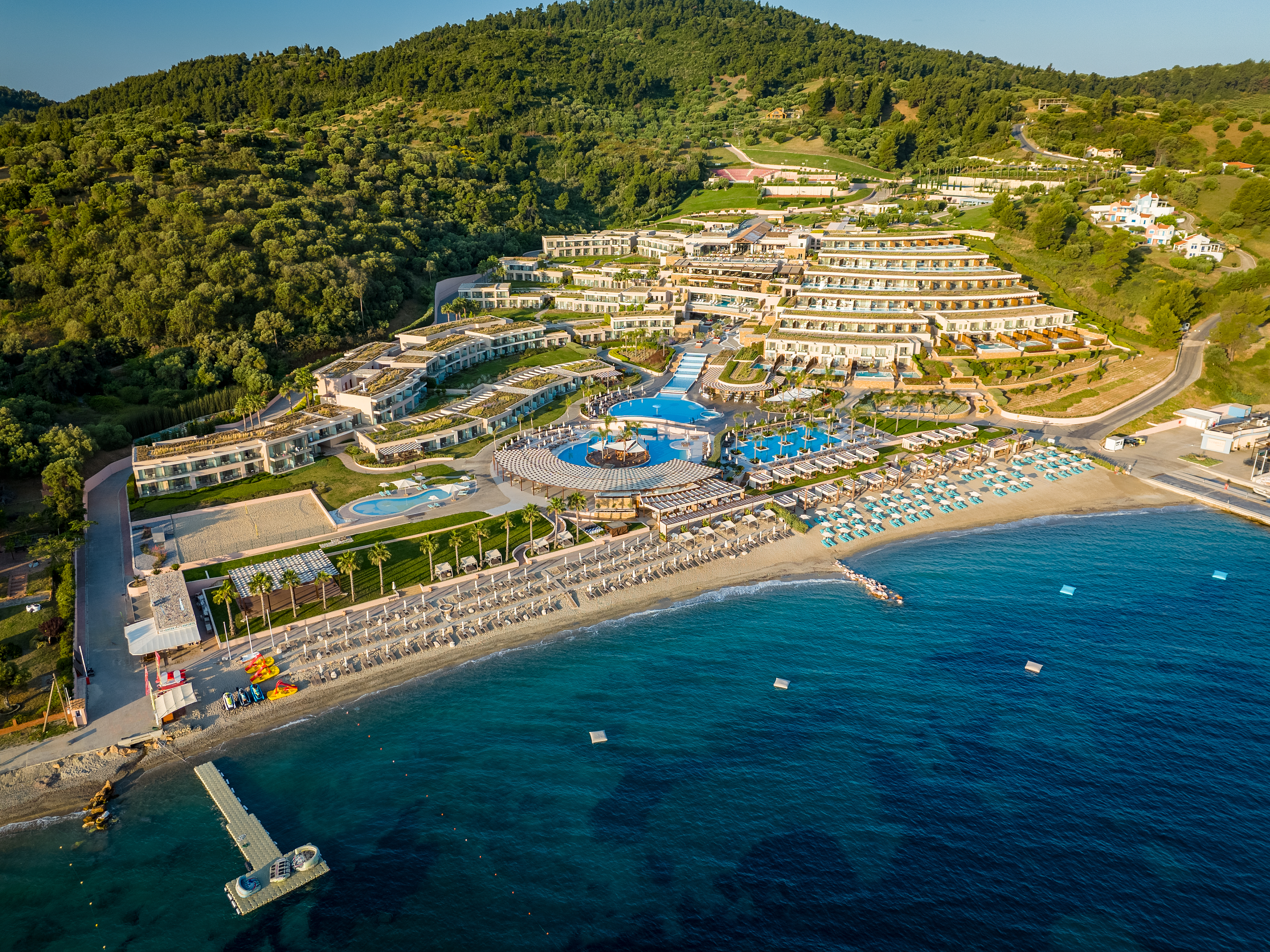 Aerial view of Miraggio Resort, with beachfront pools, turquoise umbrellas, and lush green hills surrounding the property