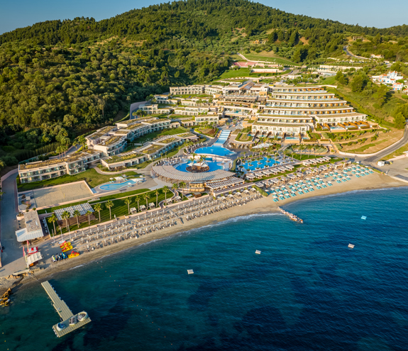 Aerial view of Miraggio Resort, with beachfront pools, turquoise umbrellas, and lush green hills surrounding the property