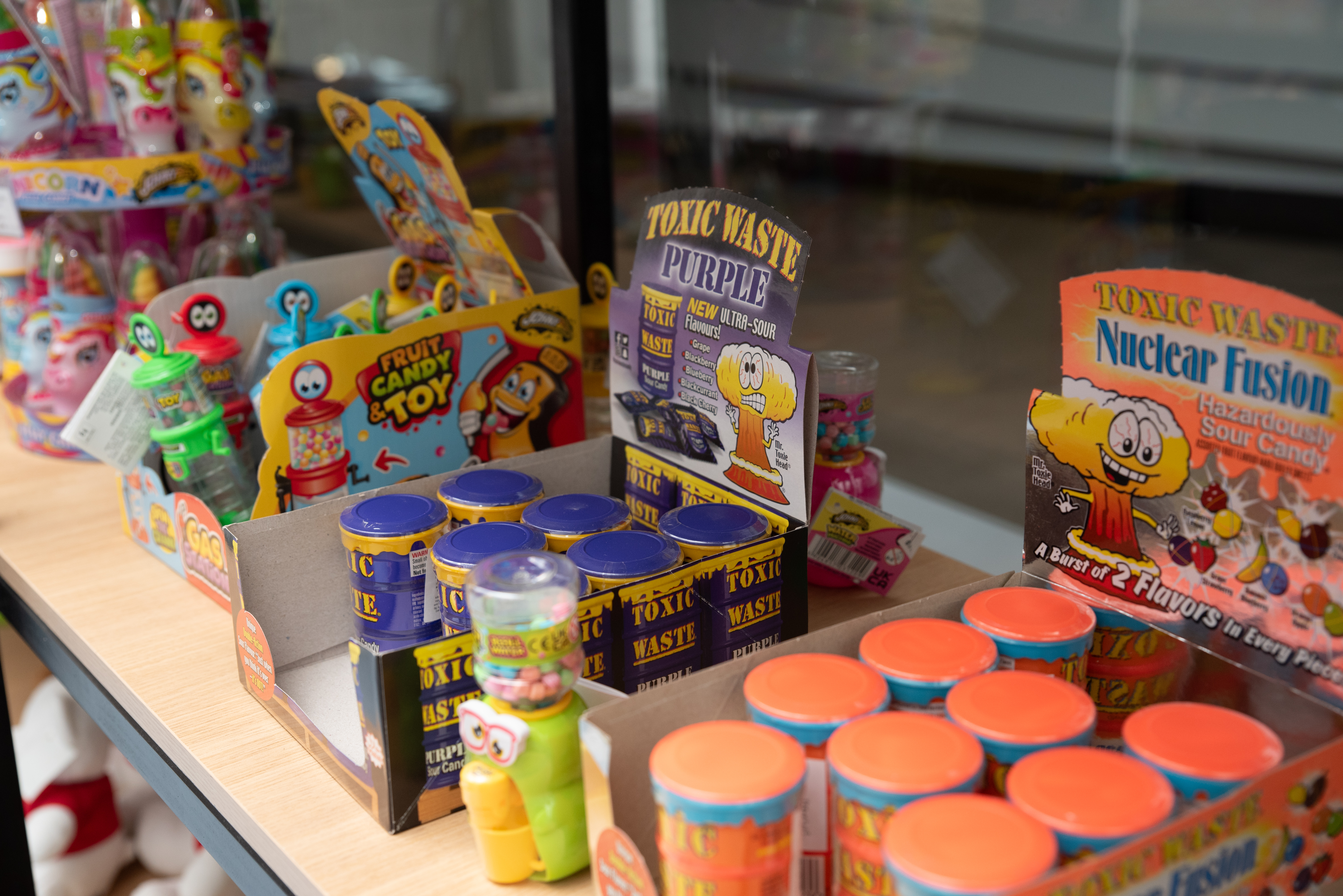 assorted novelty candies including Toxic Waste sour candy tubs and toy-shaped sweets on a wooden shelf