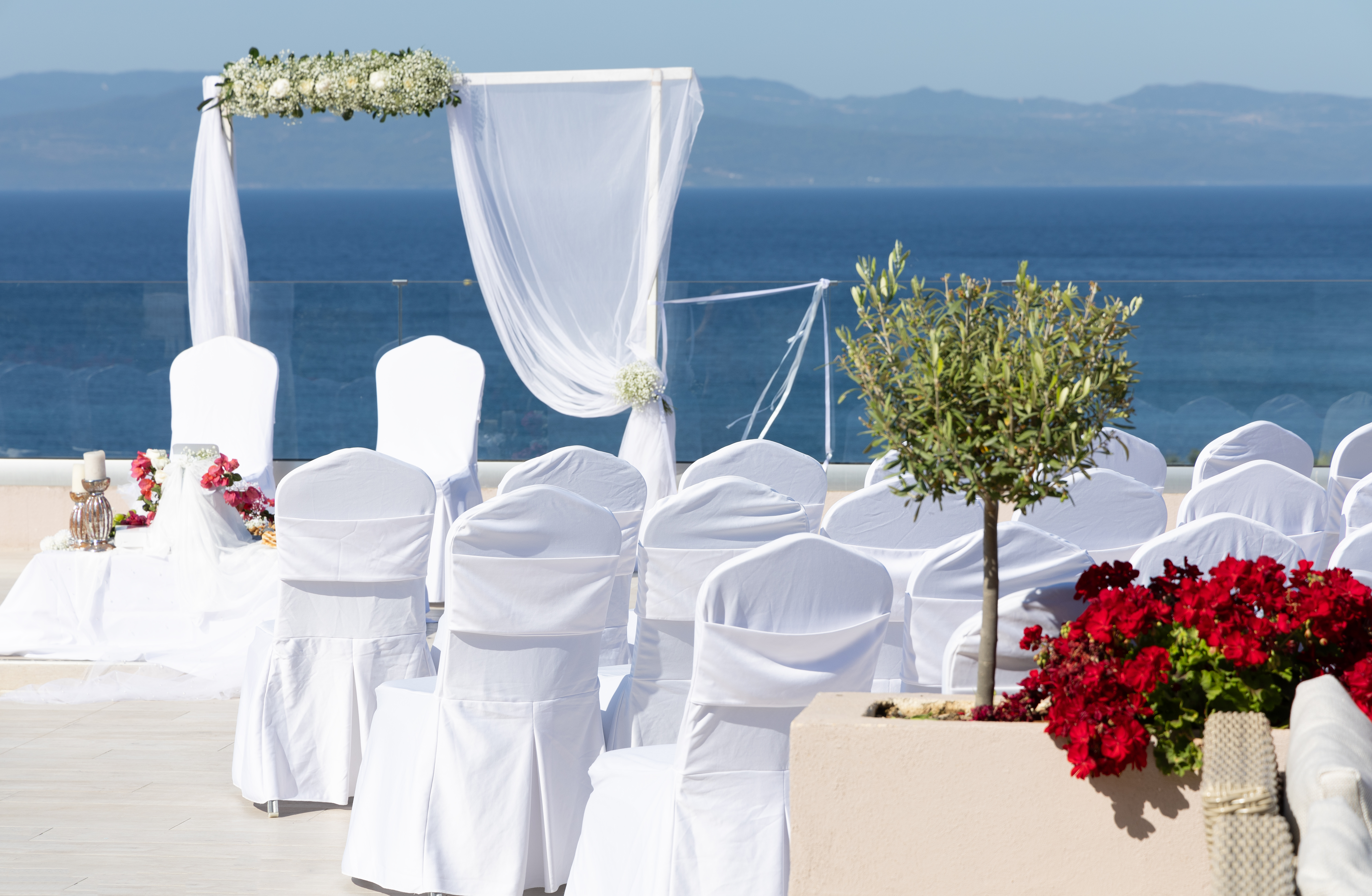Seaside wedding ceremony setup with white chairs, floral arch, and red flowers in foreground