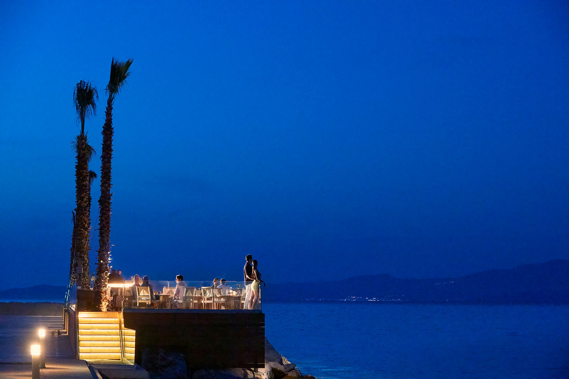Romantic couple standing at the corner of the illuminated dock, gazing at the sea under the night sky