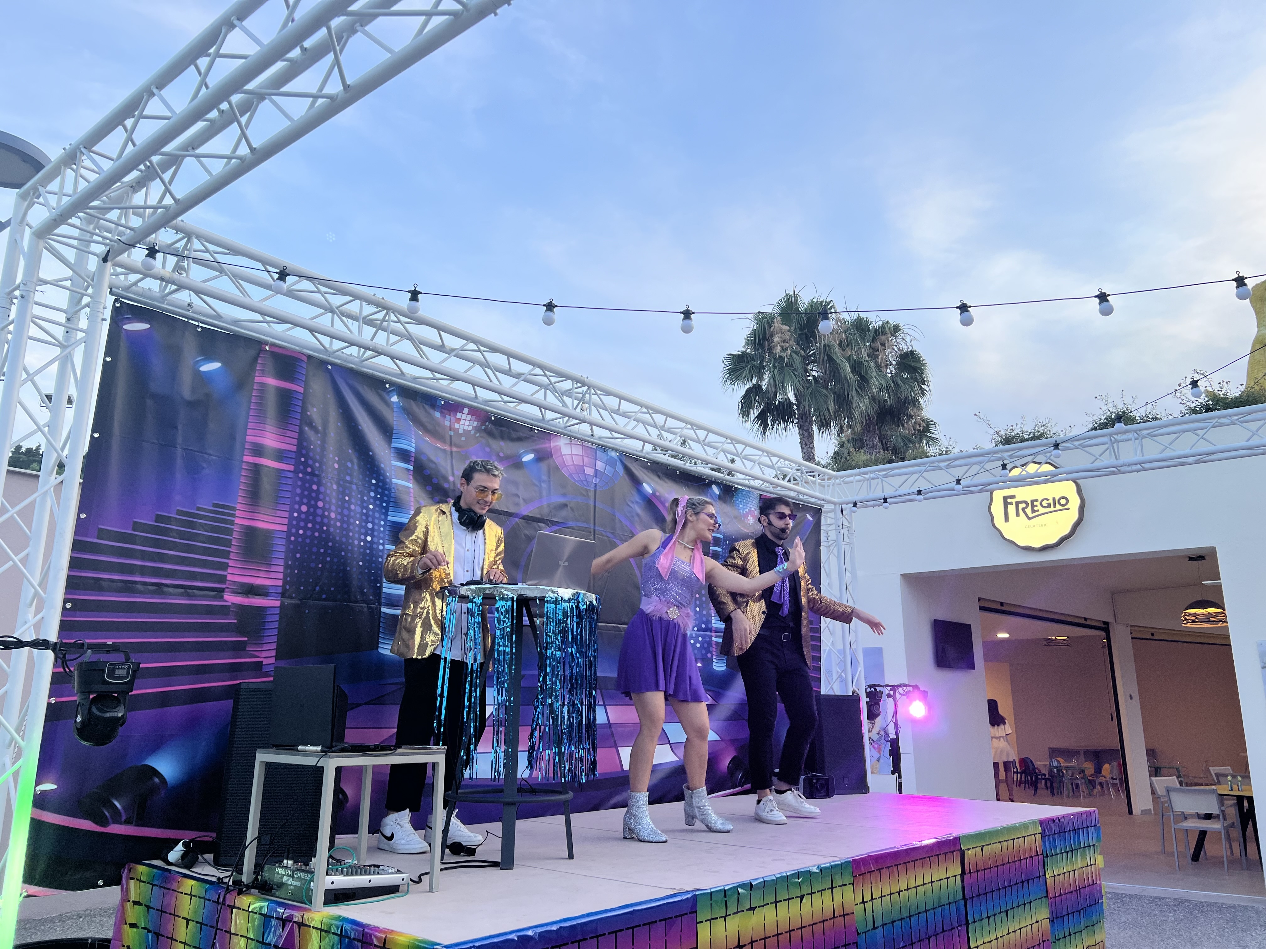 Performers in disco outfits dancing on colorful outdoor stage near Fregio cafe