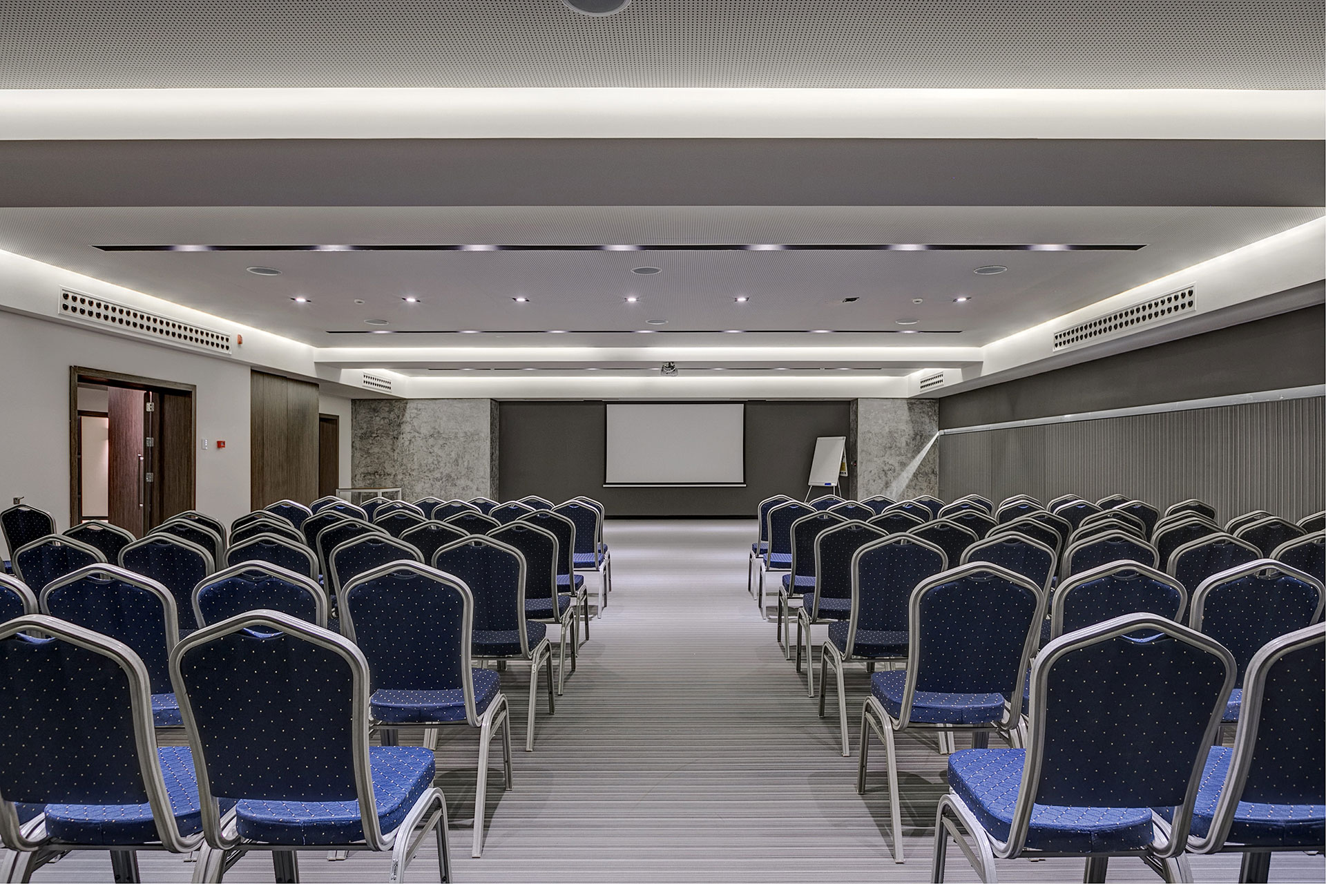 Modern conference room with rows of blue chairs, projector screen, and minimalist lighting
