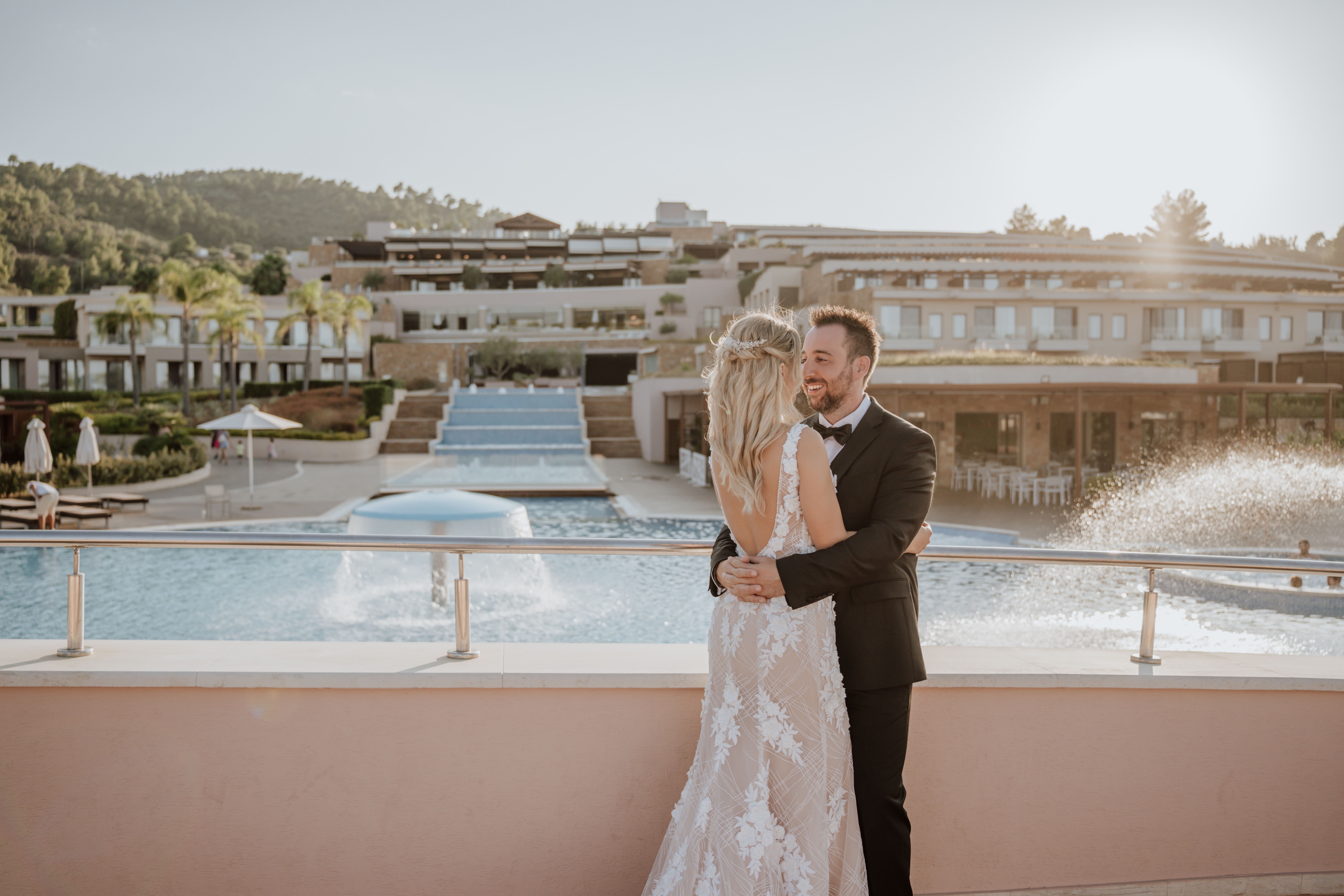 Bride and groom embracing by the pool with cascading water and resort view at sunset