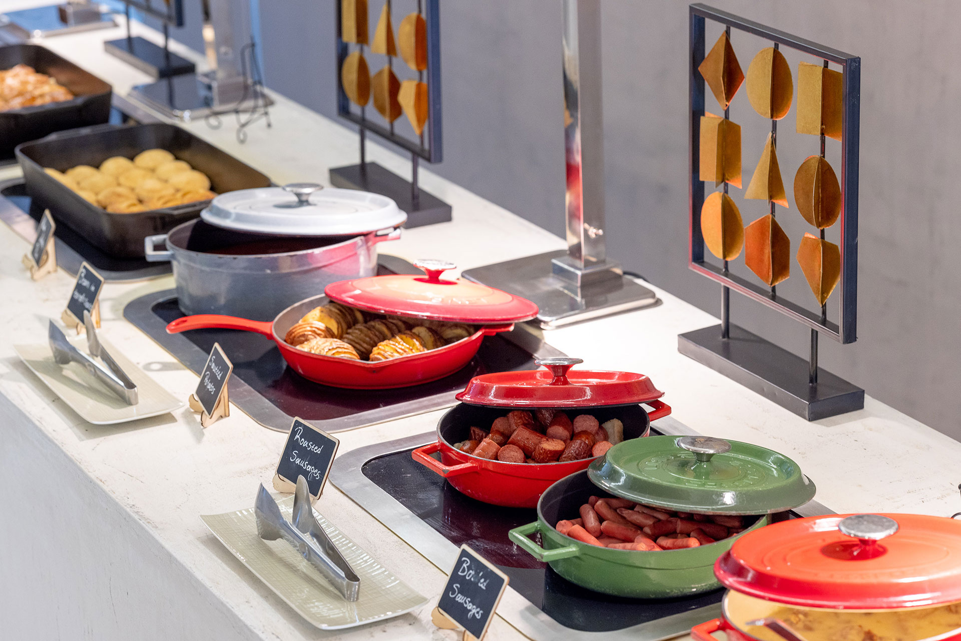 Hot breakfast buffet with roasted and boiled sausages, potatoes, and pastries in colorful cast-iron pots