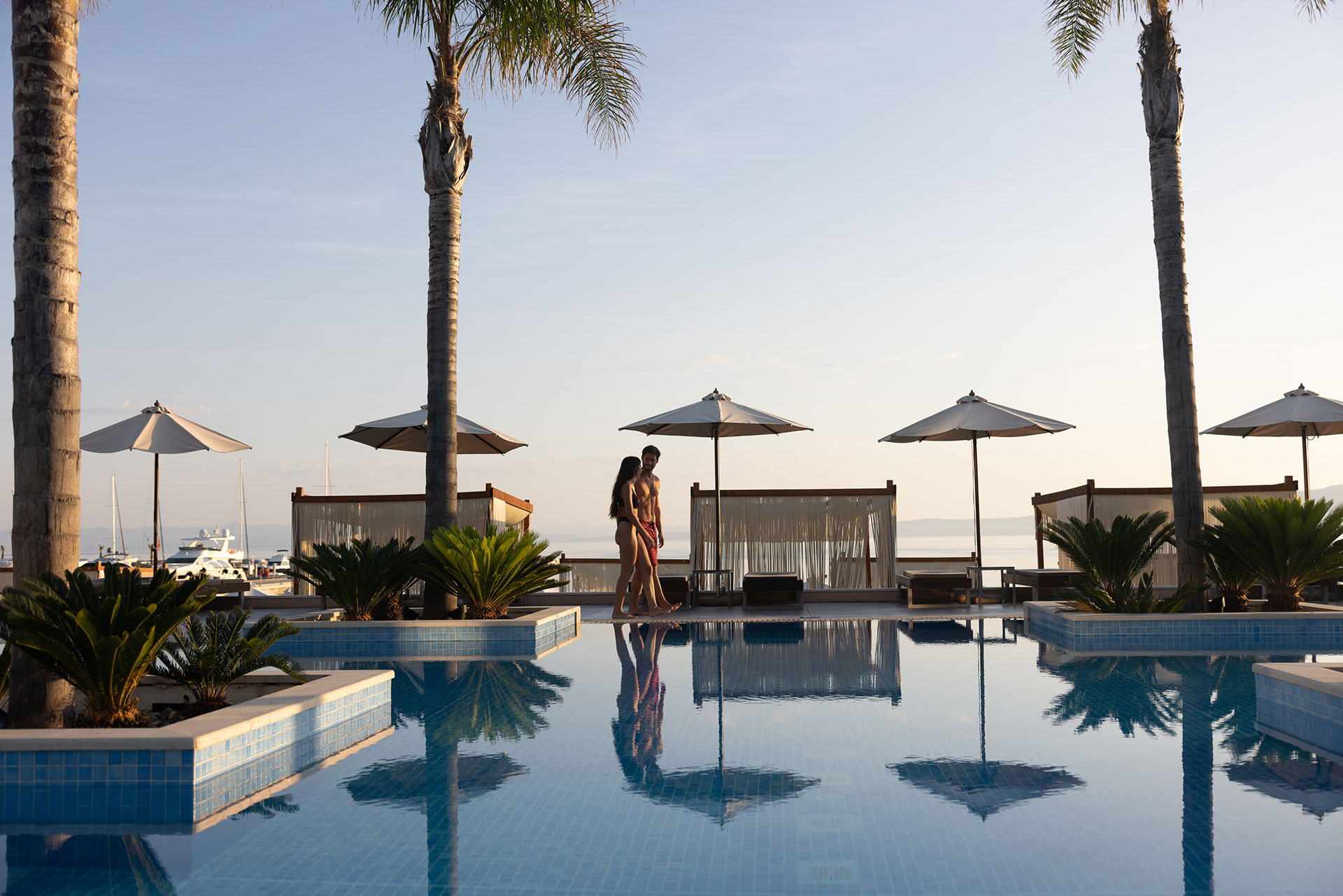 Couple walking by the serene pool surrounded by cabanas and palm trees at sunrise