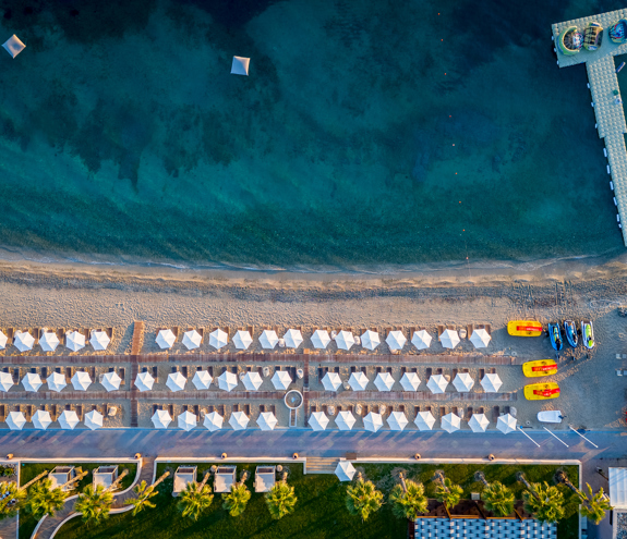 Top down aerial view of the sandy beach with aligned umbrellas, turquoise waters, and a pier with colorful water sports equipment