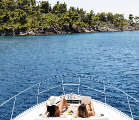 Two women sunbathing and relaxing on the bow of a yacht anchored near a lush green coastline