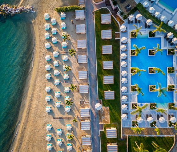 Aerial view of the beachfront and pool area, showcasing turquoise waters, sunbeds and umbrellas, and cabanas