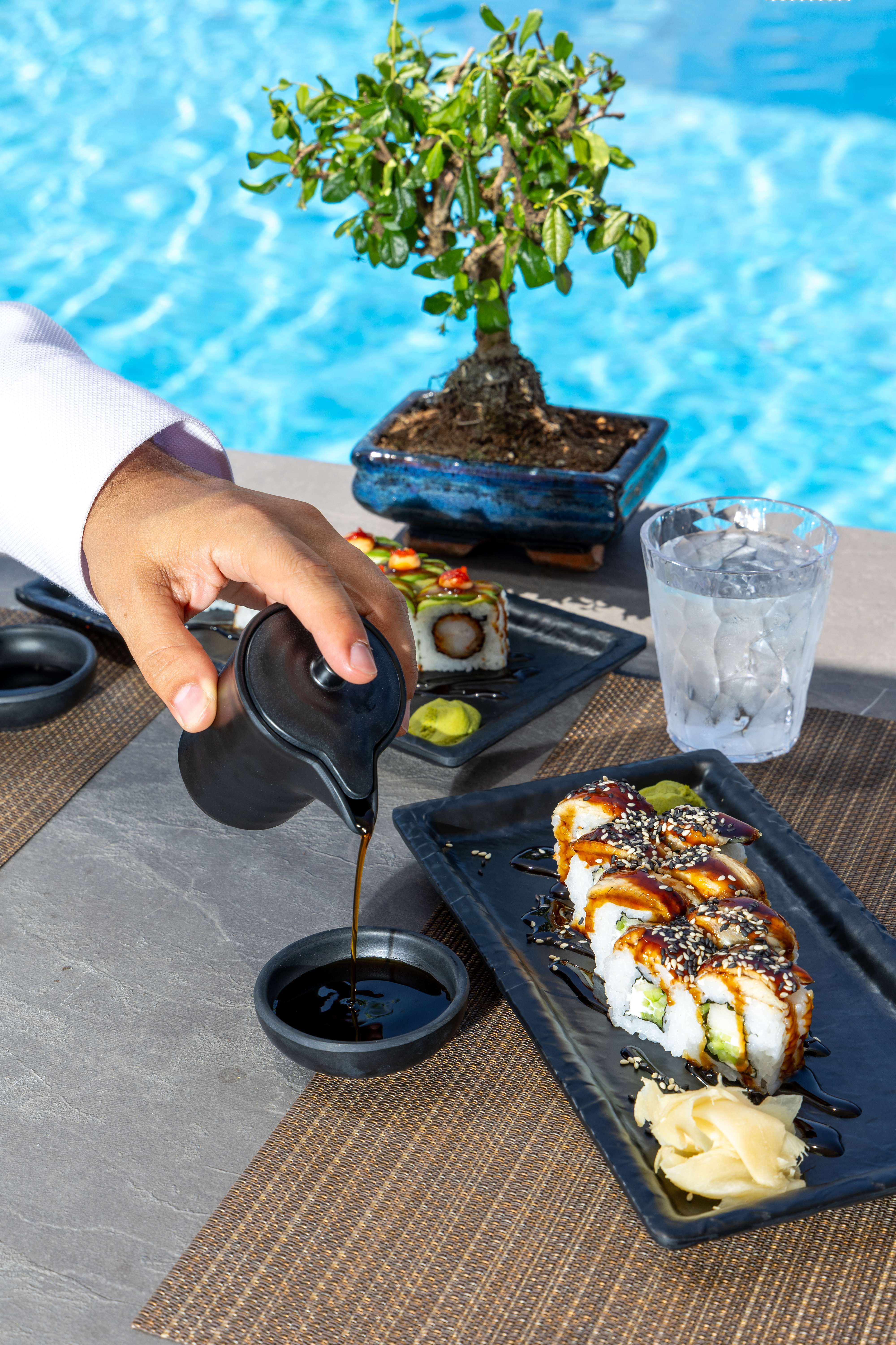 Hand pouring sauce beside sushi rolls and bonsai tree by the pool