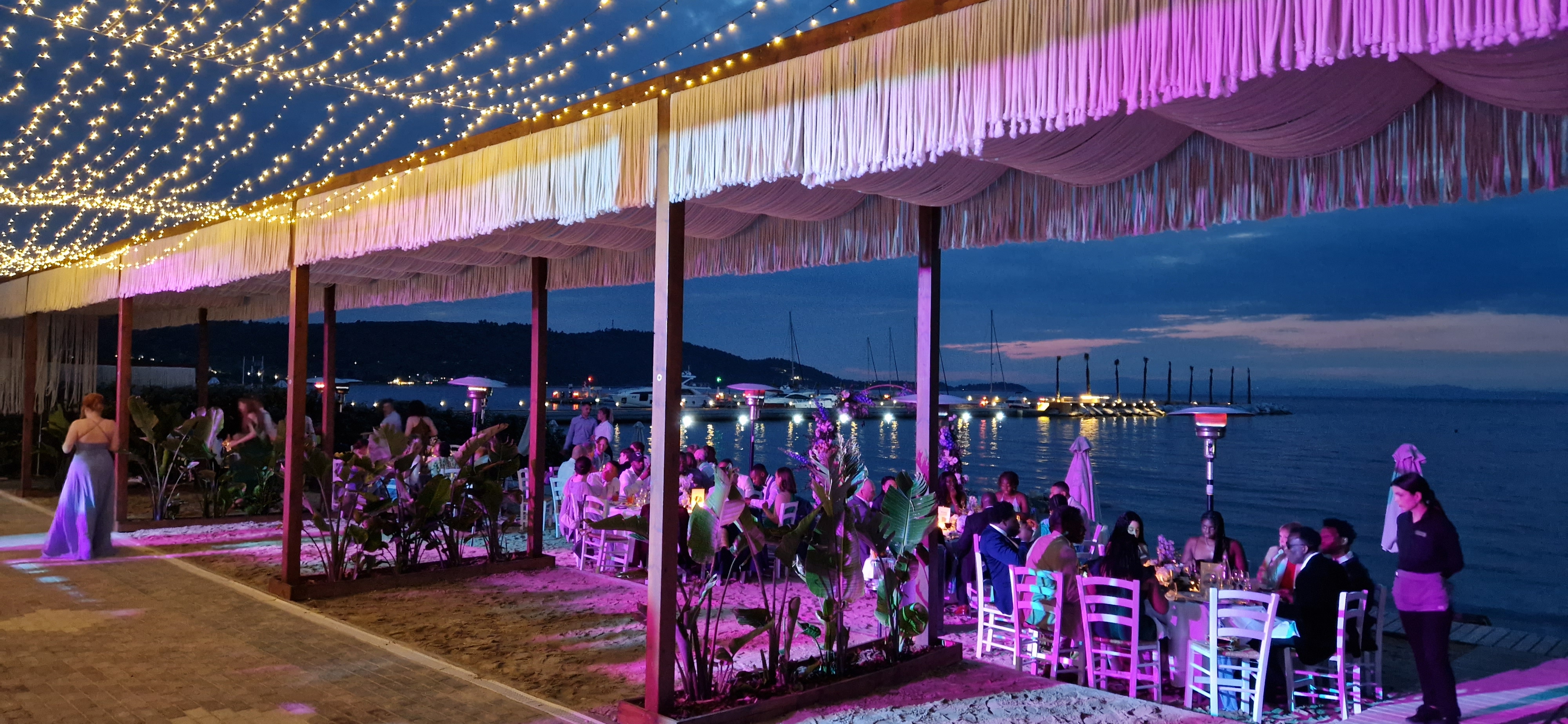 Evening beach wedding reception under fairy lights with sea and yachts