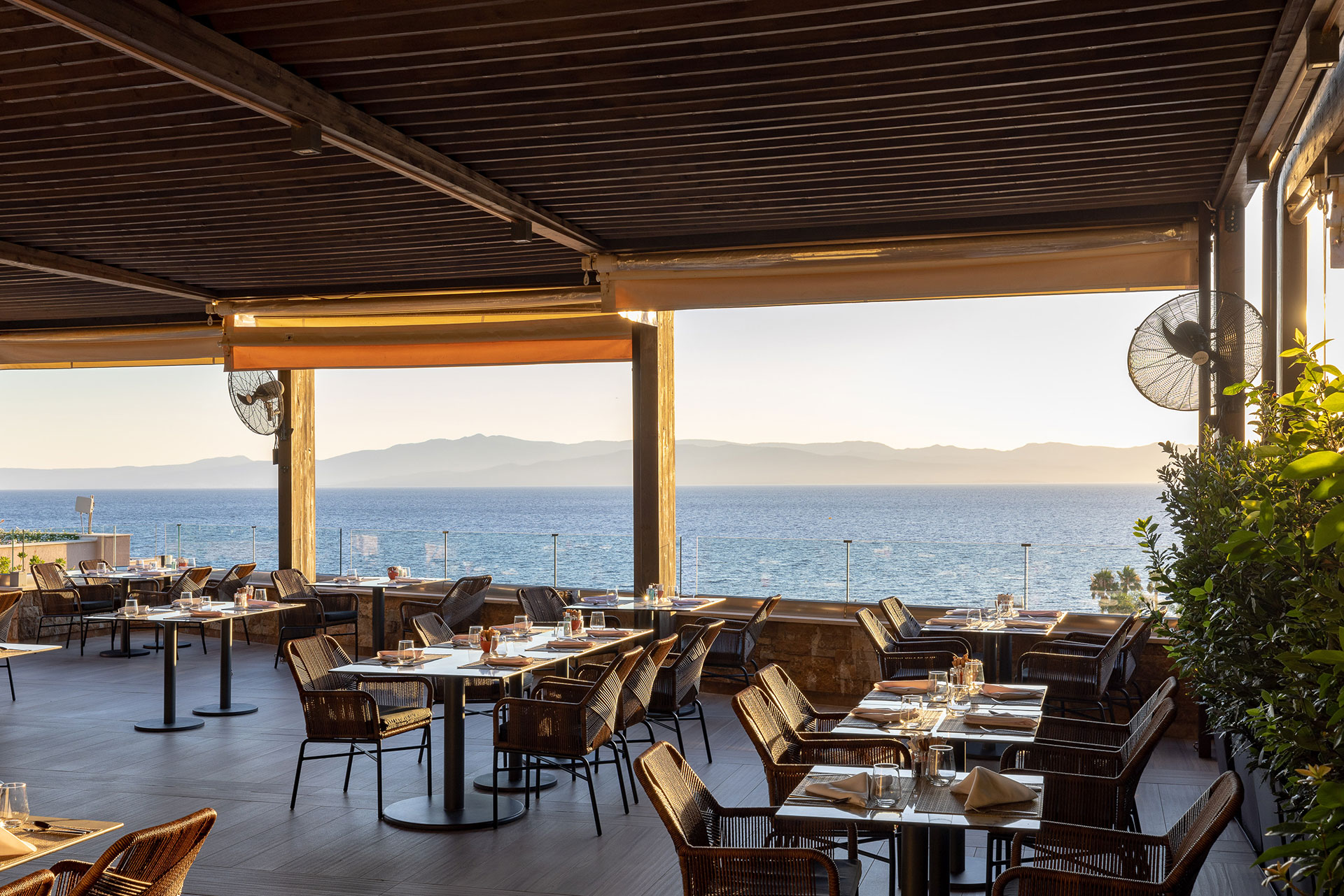 Outdoor terrace of Elia Restaurant overlooking the Aegean Sea at sunset, with elegant tables and rattan chairs