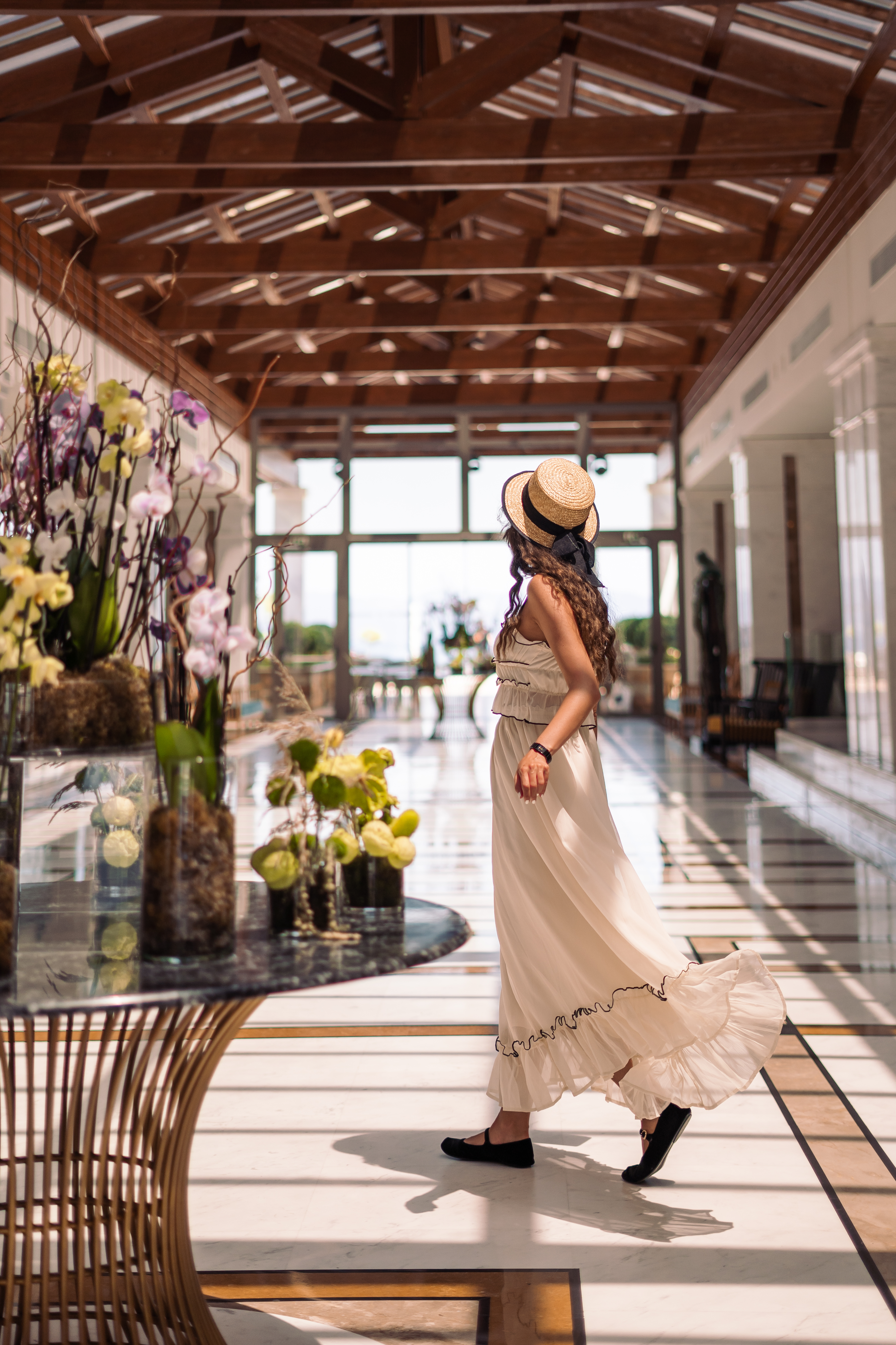 Woman in flowing beige dress and straw hat walking through the elegant marble lobby with floral decor
