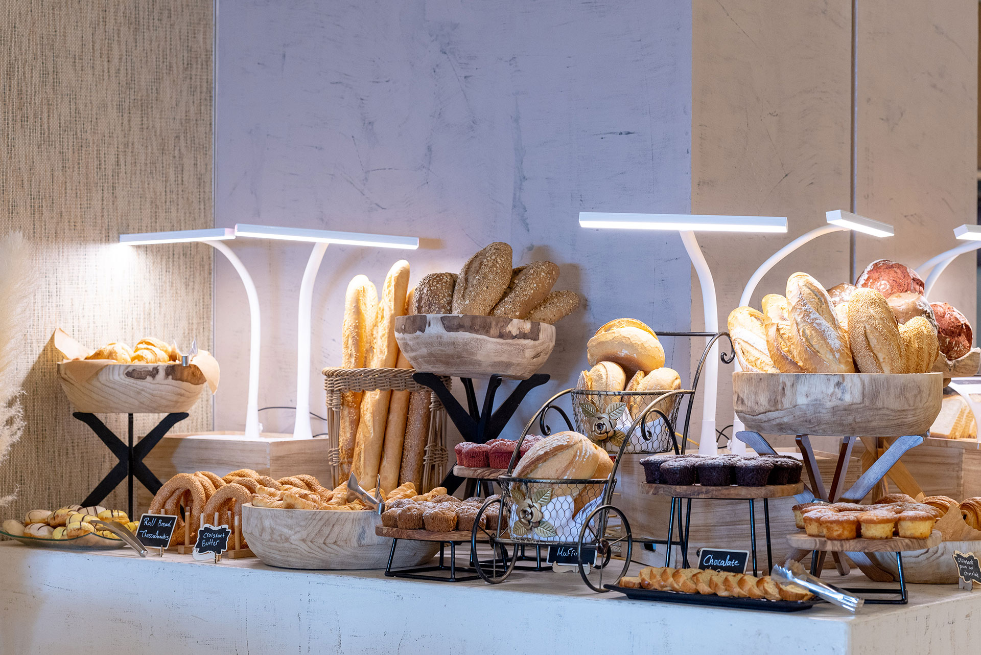 Assorted breads and pastries including baguettes, muffins, and croissants displayed under warm lighting