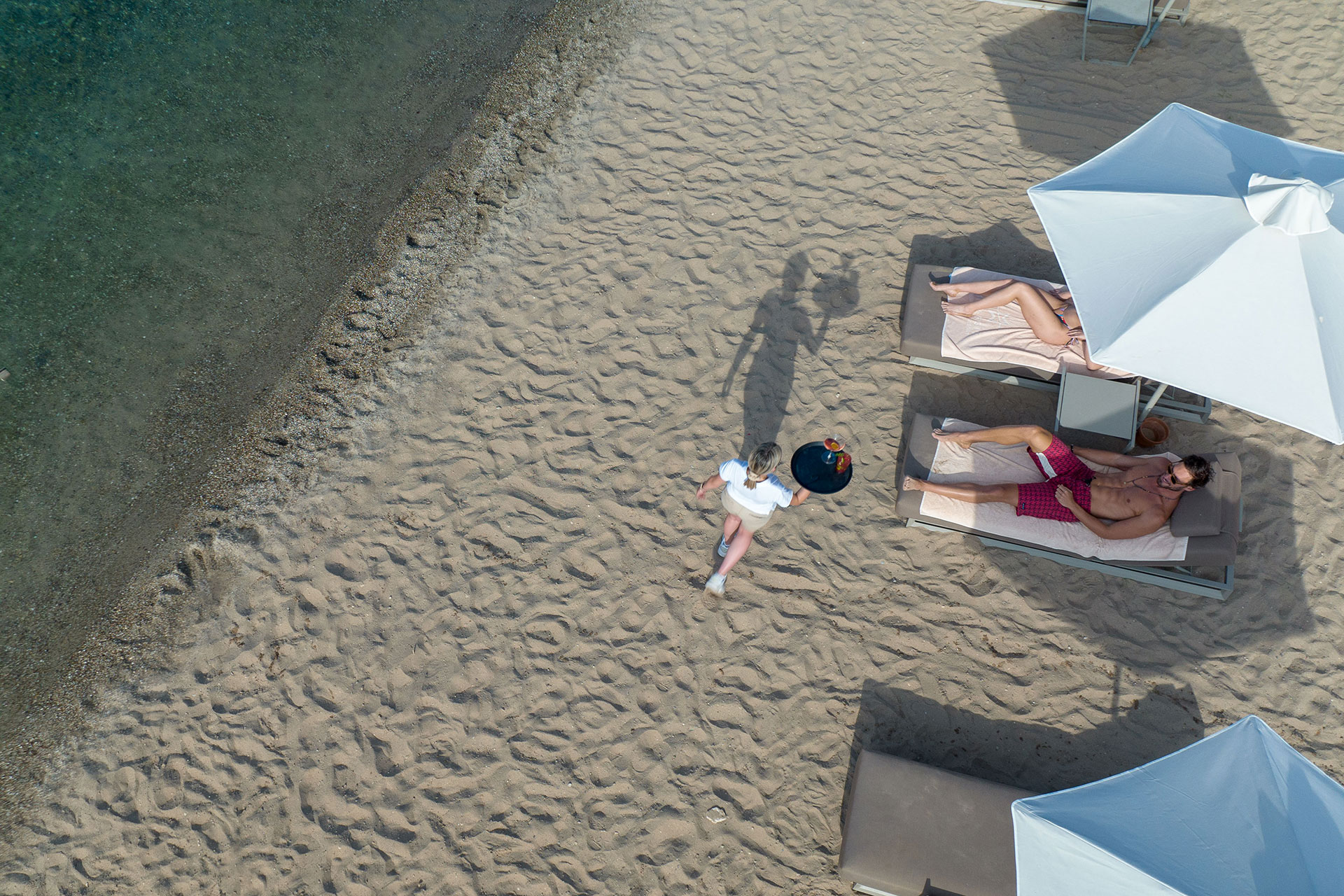 Top down view of a waitress serving drinks to a couple enjoying the sandy beach and clear blue sea