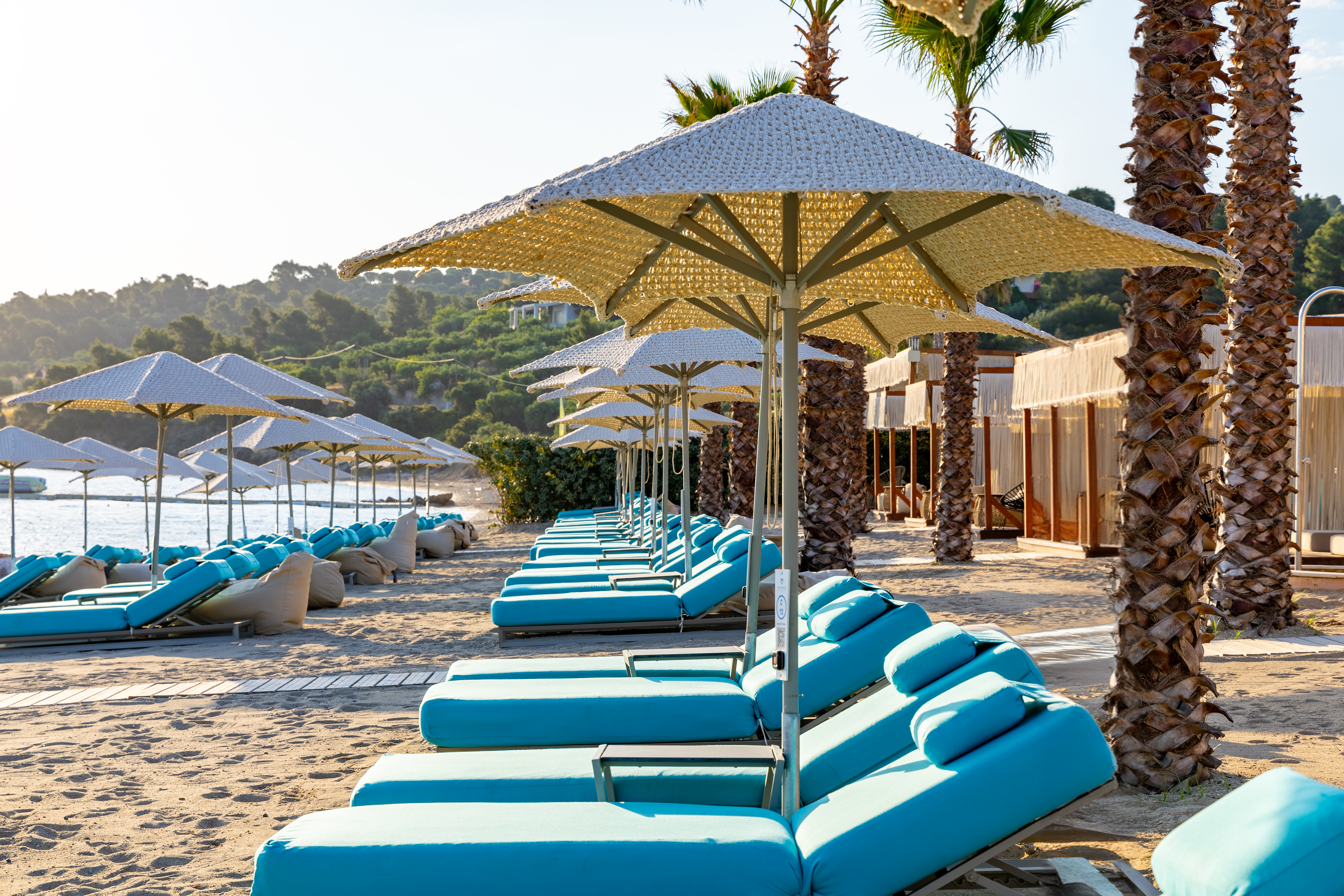 Row of turquoise sunbeds under woven umbrellas along the palm-lined sandy beach in afternoon light