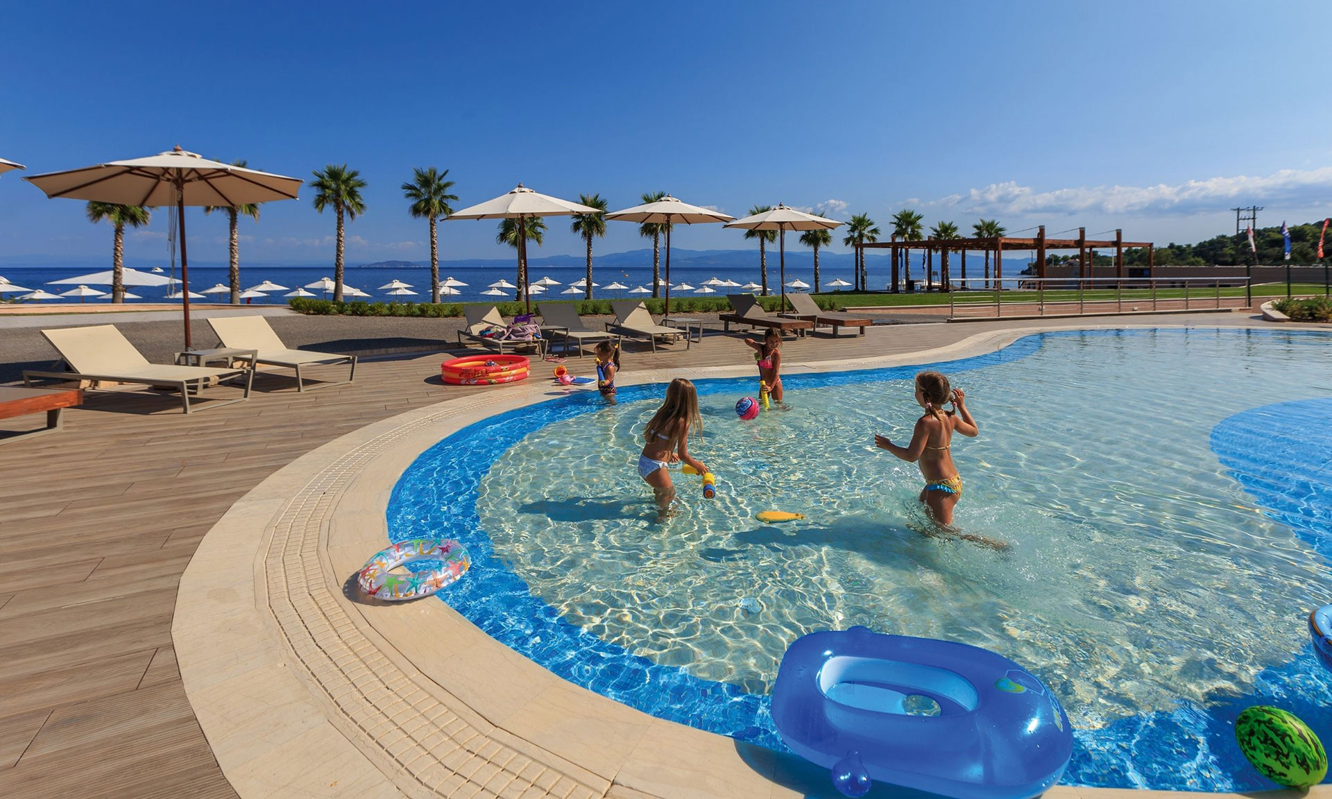 Children playing in a shallow outdoor pool with sea view and palm trees